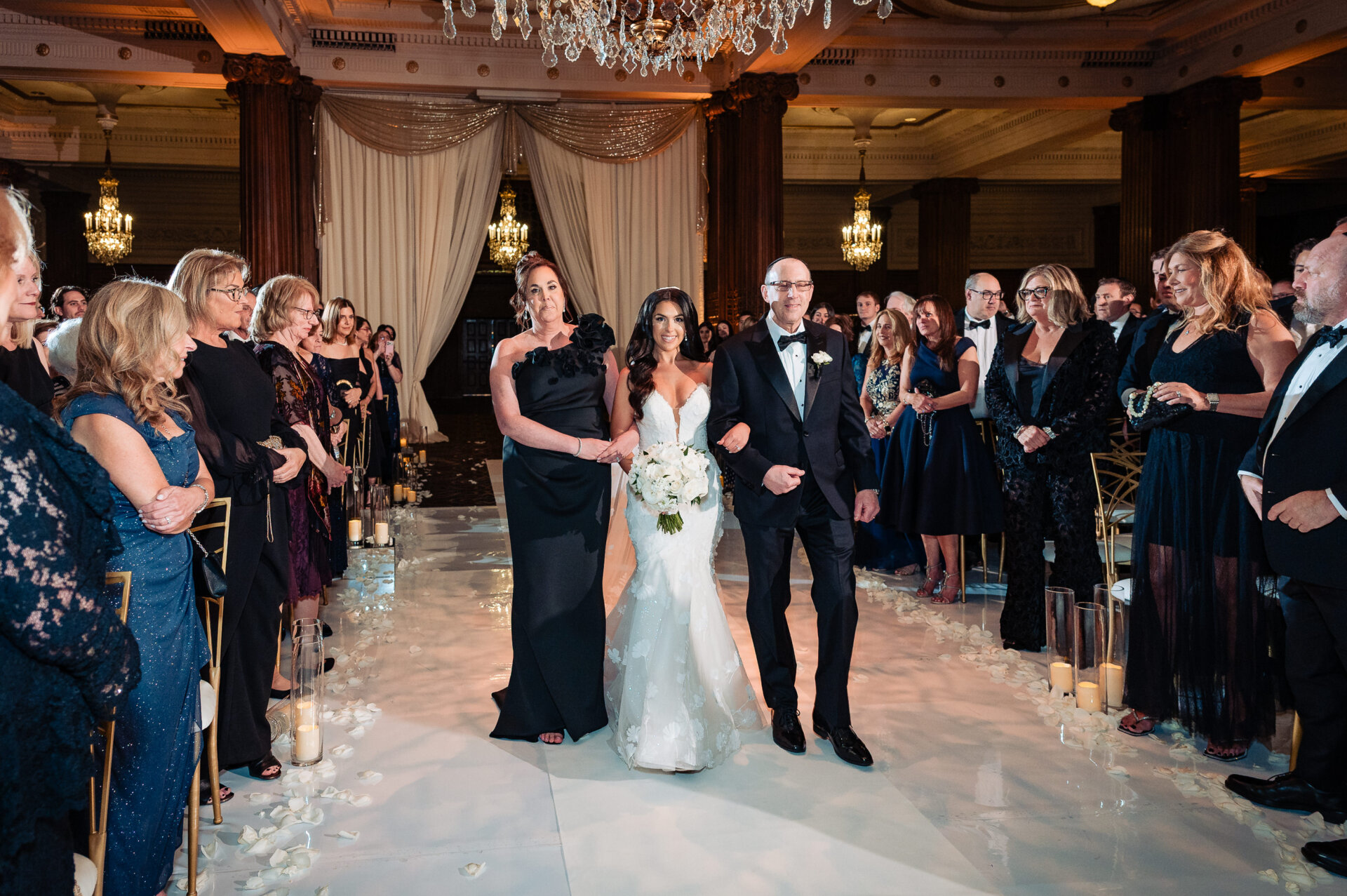 The bride being escorted down a white aisle by her parents in a grand ballroom under crystal chandeliers.