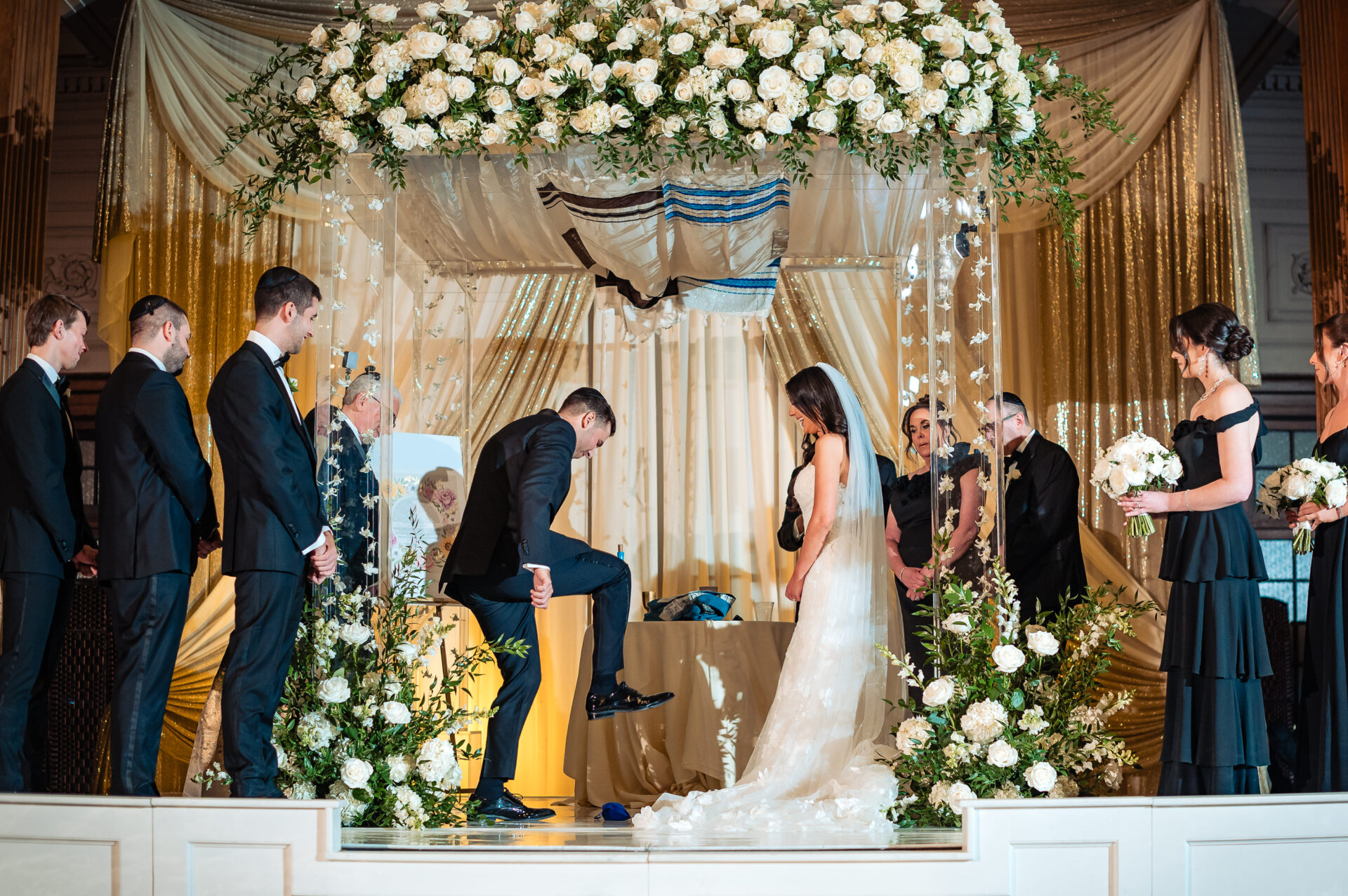 The groom breaking a glass during the Jewish wedding ceremony while the bride watches. This traditional moment takes place under an elegant chuppah decorated with lush white flowers and greenery.