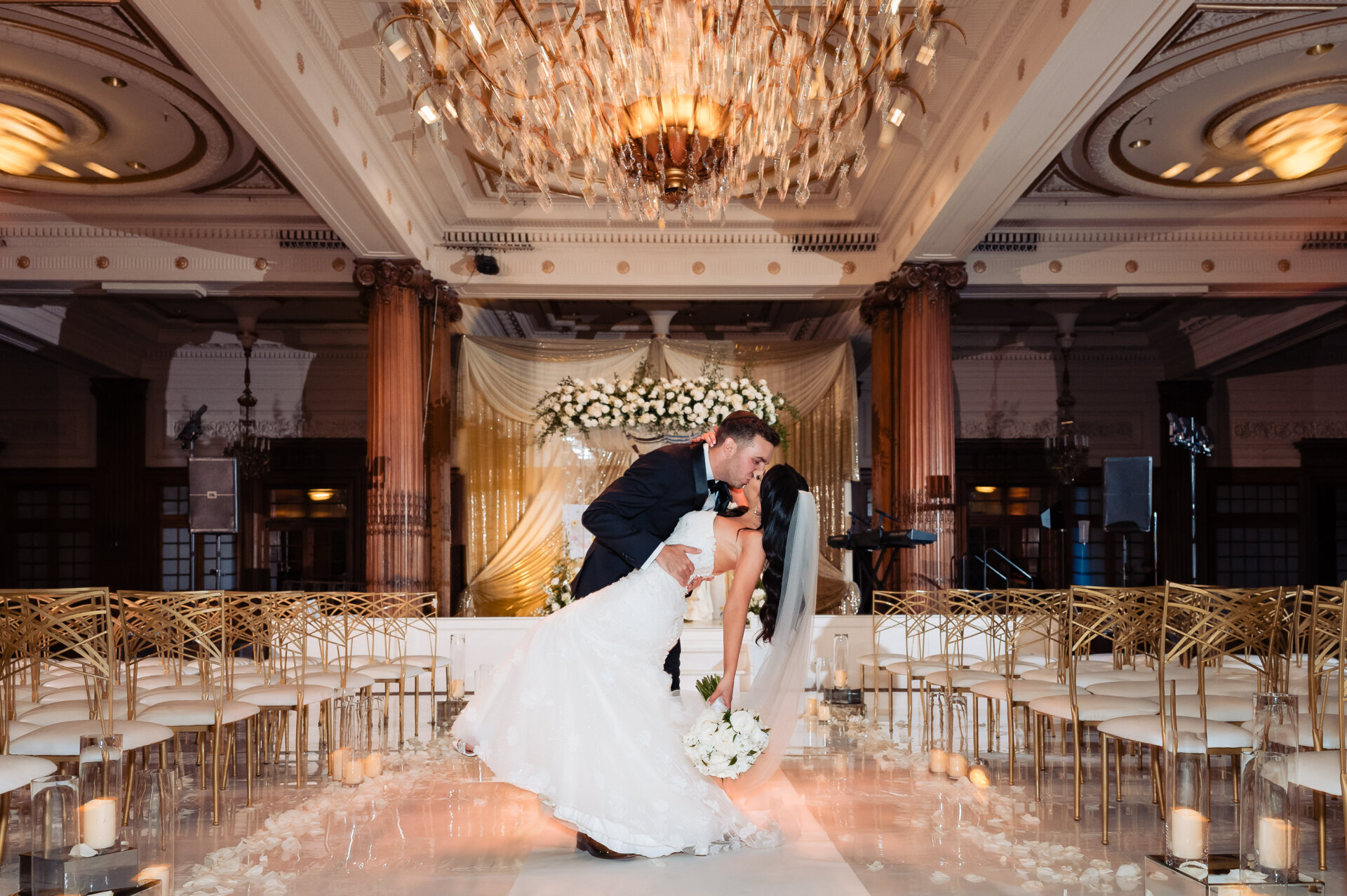 A newlywed couple shares a passionate kiss on a grand ballroom dance floor. The groom in a black tuxedo dips his bride in a white wedding gown with a flowing train and long veil.