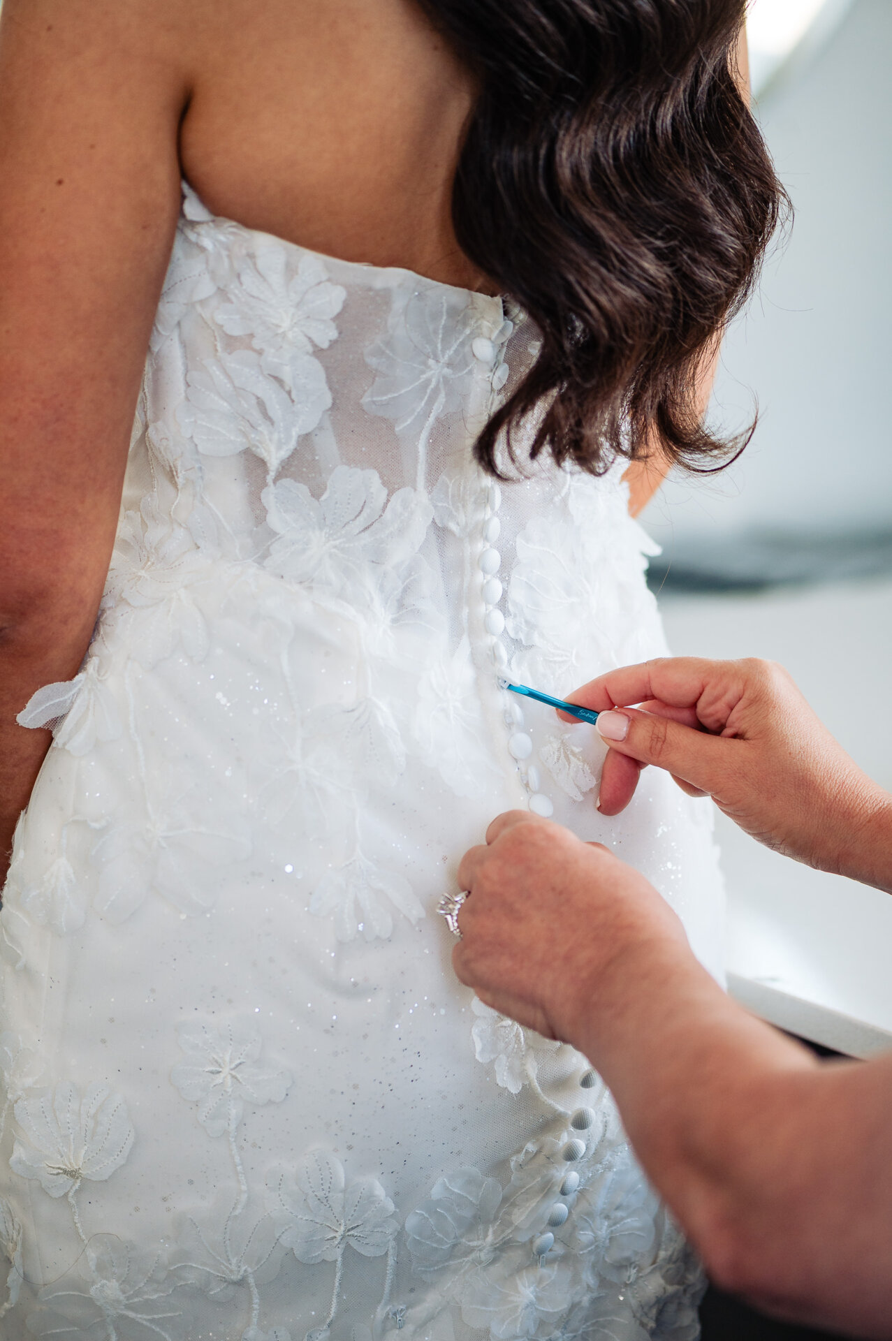 "Close-up of bridesmaid fastening buttons on back of bride's white floral applique wedding gown