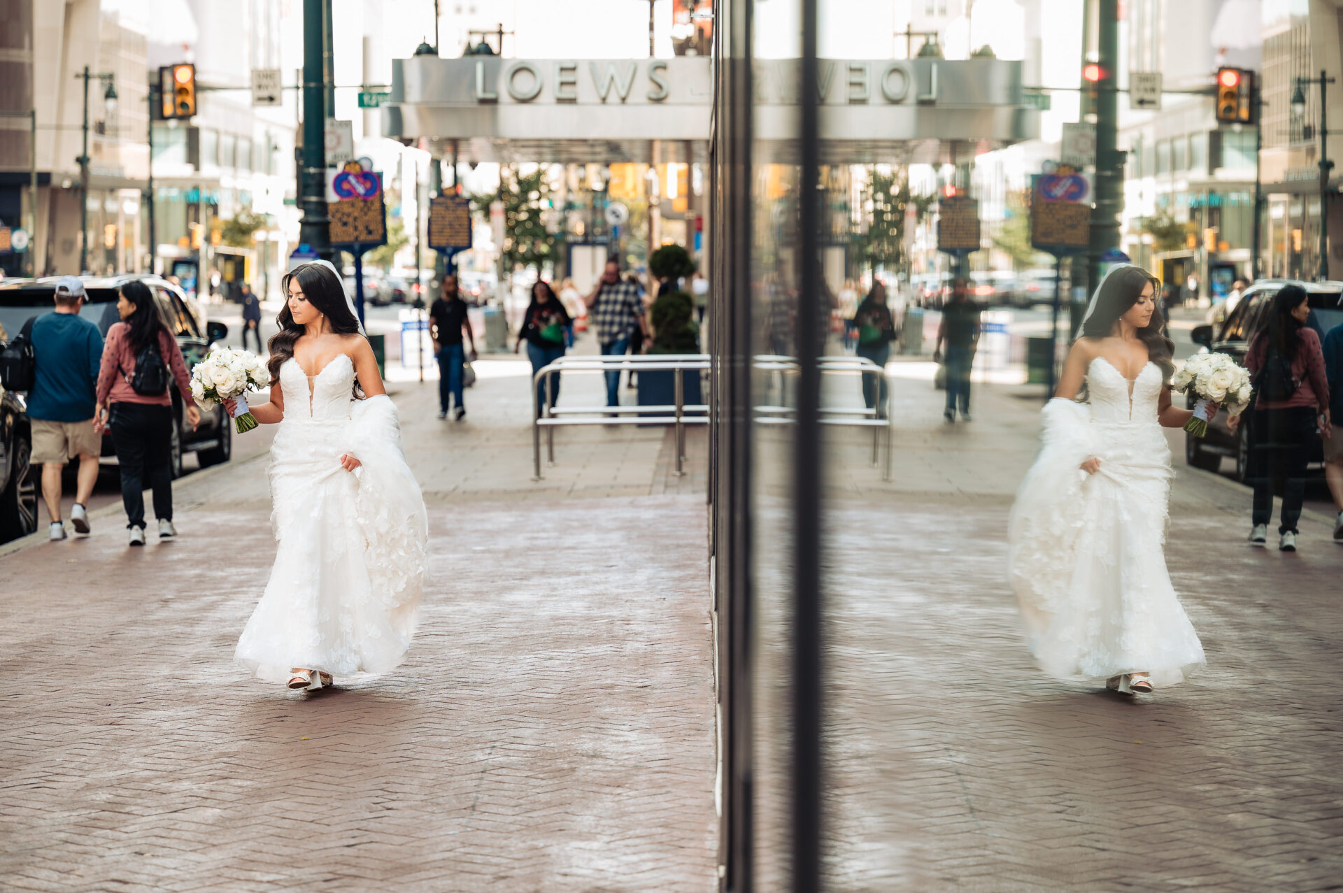 Bride in flowing white ballgown with bouquet walking on city street reflected in glass storefront near crystal tea room hotel