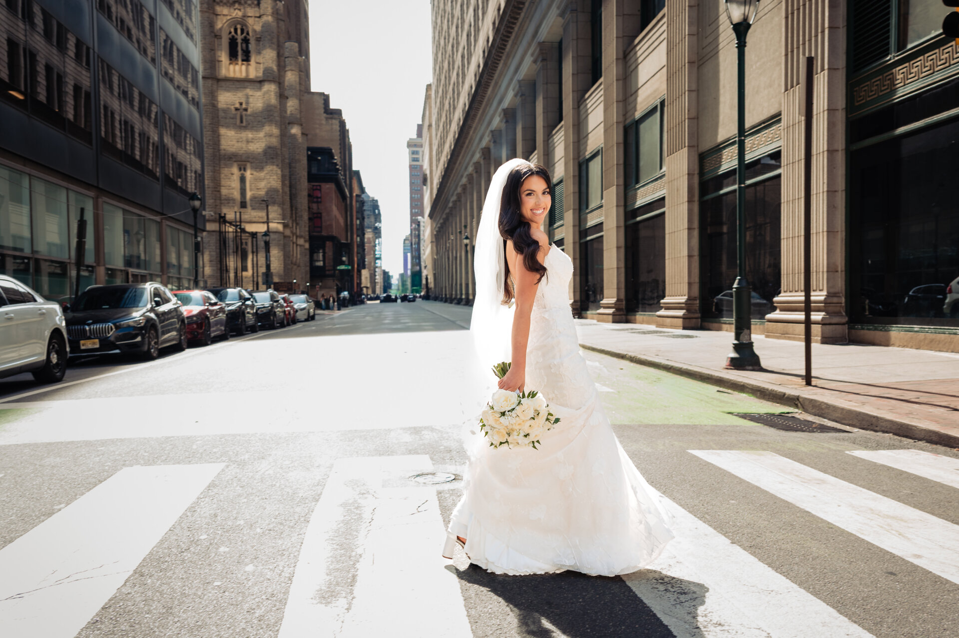 Bride in fitted white gown with cathedral veil holding white bouquet standing in middle of city crosswalk with historic church tower in background