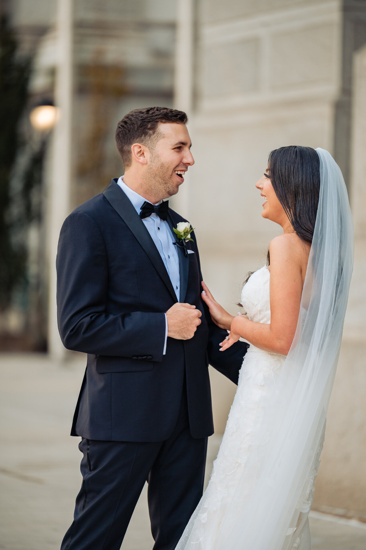 Newlyweds laughing together in first look moment, groom in navy tuxedo and bride in strapless white gown with veil