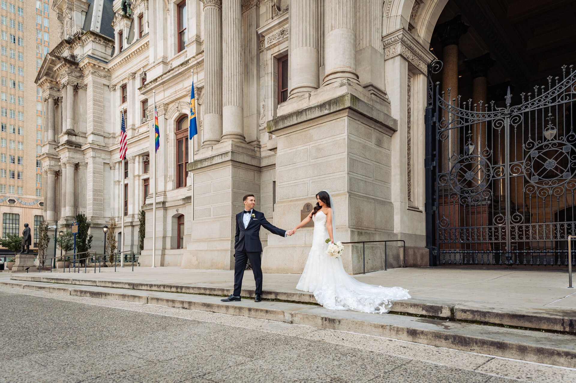 Newlywed couple holding hands in front of the ornate architecture and iron gates of Philadelphia City Hall