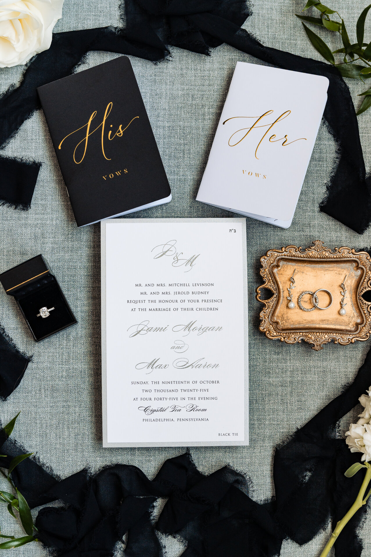 Wedding flatlay with His and Her vow books in gold calligraphy, formal invitation, engagement ring in black box, and wedding rings on ornate gold tray with black silk ribbon and white roses