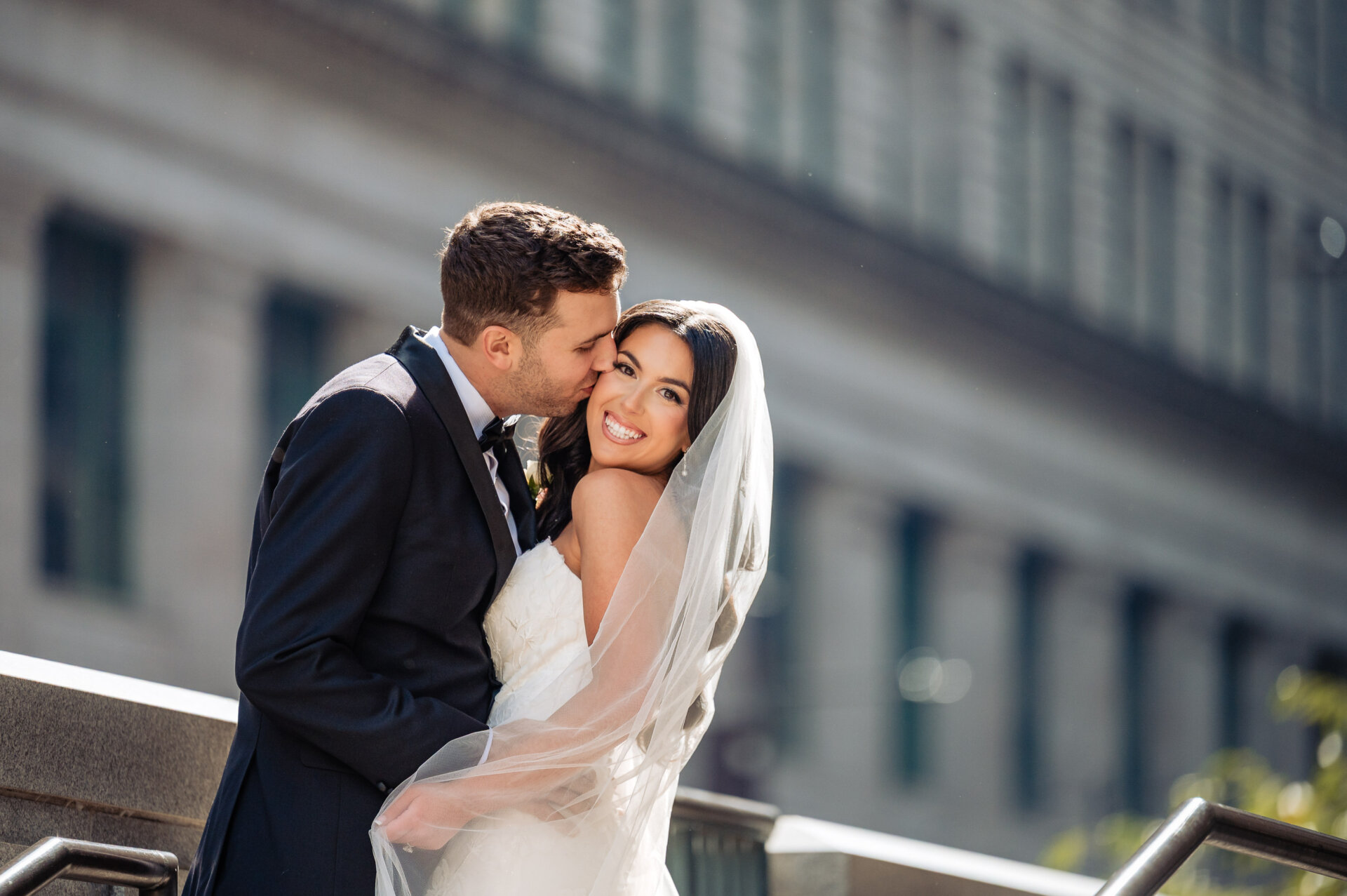 Groom kissing the bride on the forehead as she smiles at the camera, featuring a blurred city building in the background.