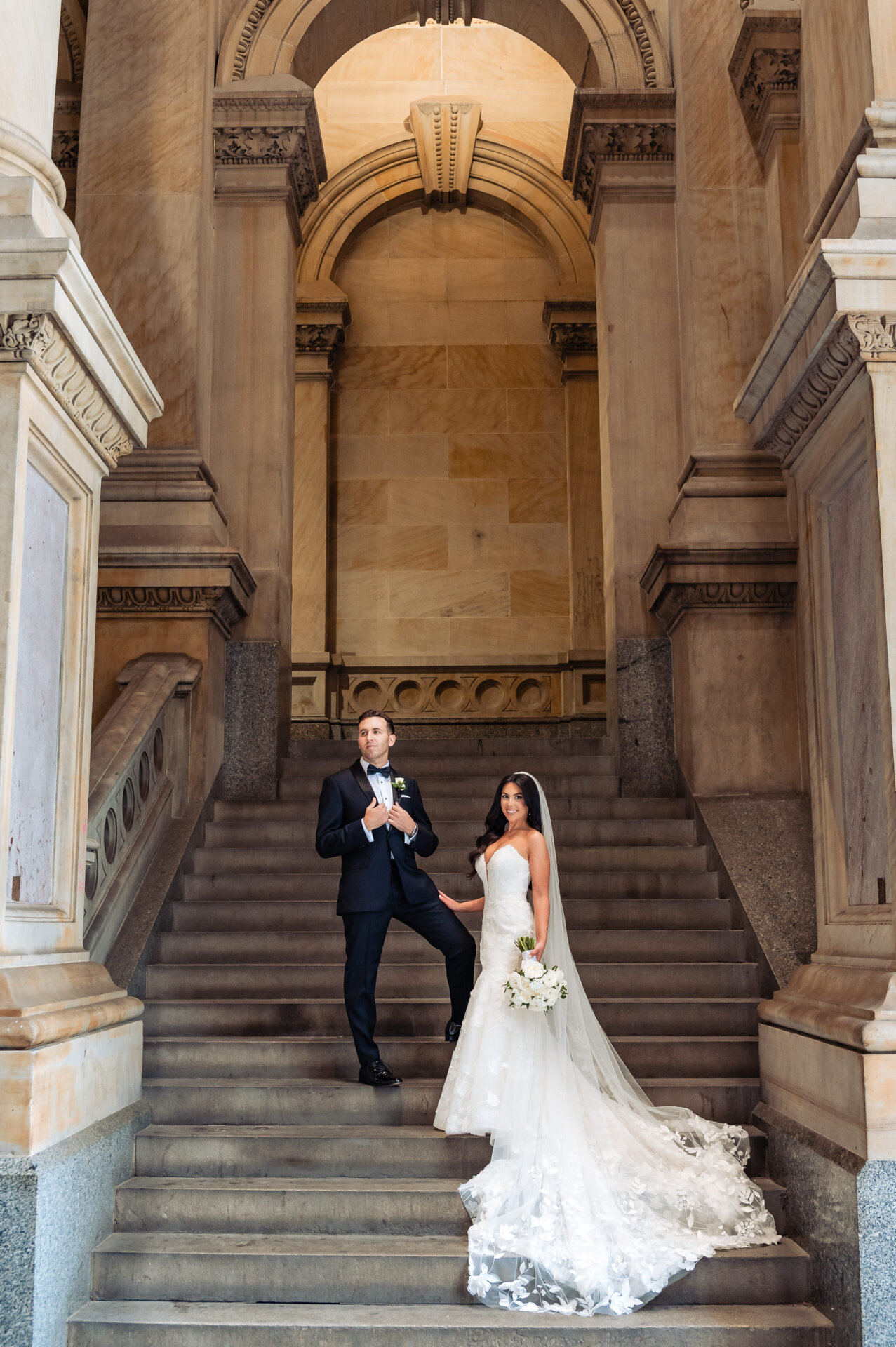 Bride and groom on marble staircase under ornate arched colonnade with dramatic cathedral train displayed on steps