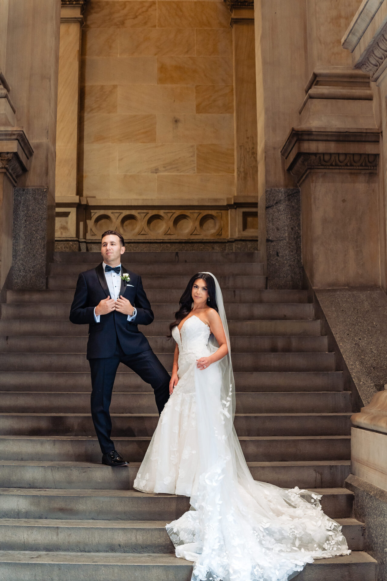 A bride and groom posing on a grand stone staircase inside a historic building with high ceilings.
