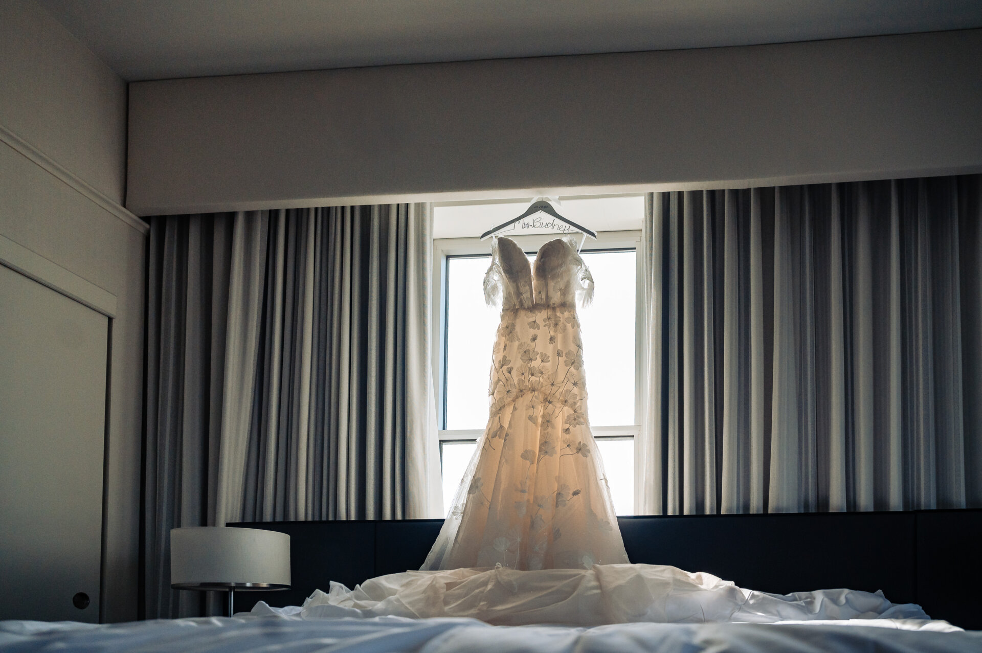 Strapless wedding gown with floral applique and flowing train hanging on personalized hanger in window of hotel room