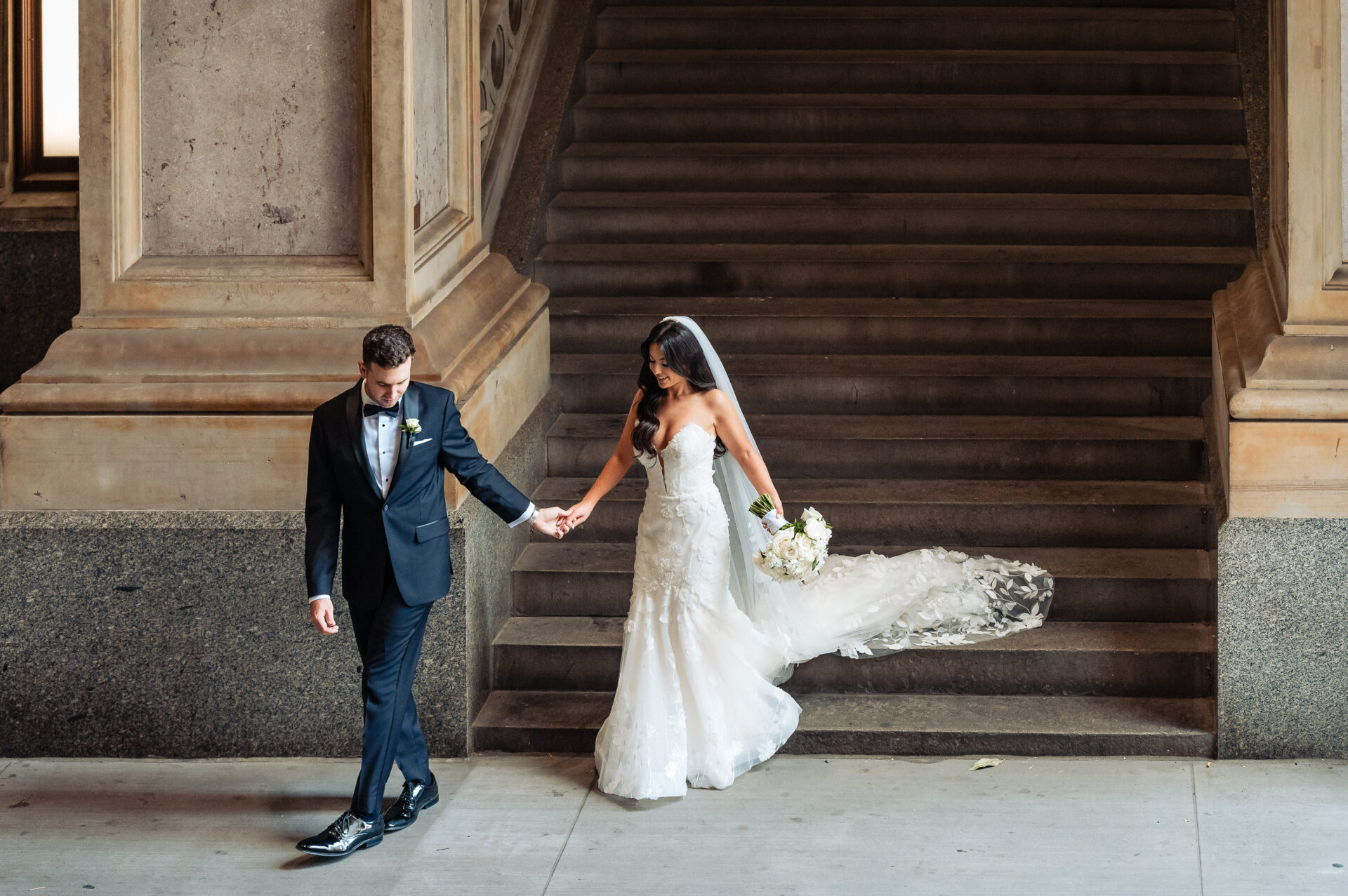 Groom in navy tuxedo leading bride in white gown with flowing train down grand stone staircase under ornate archway
