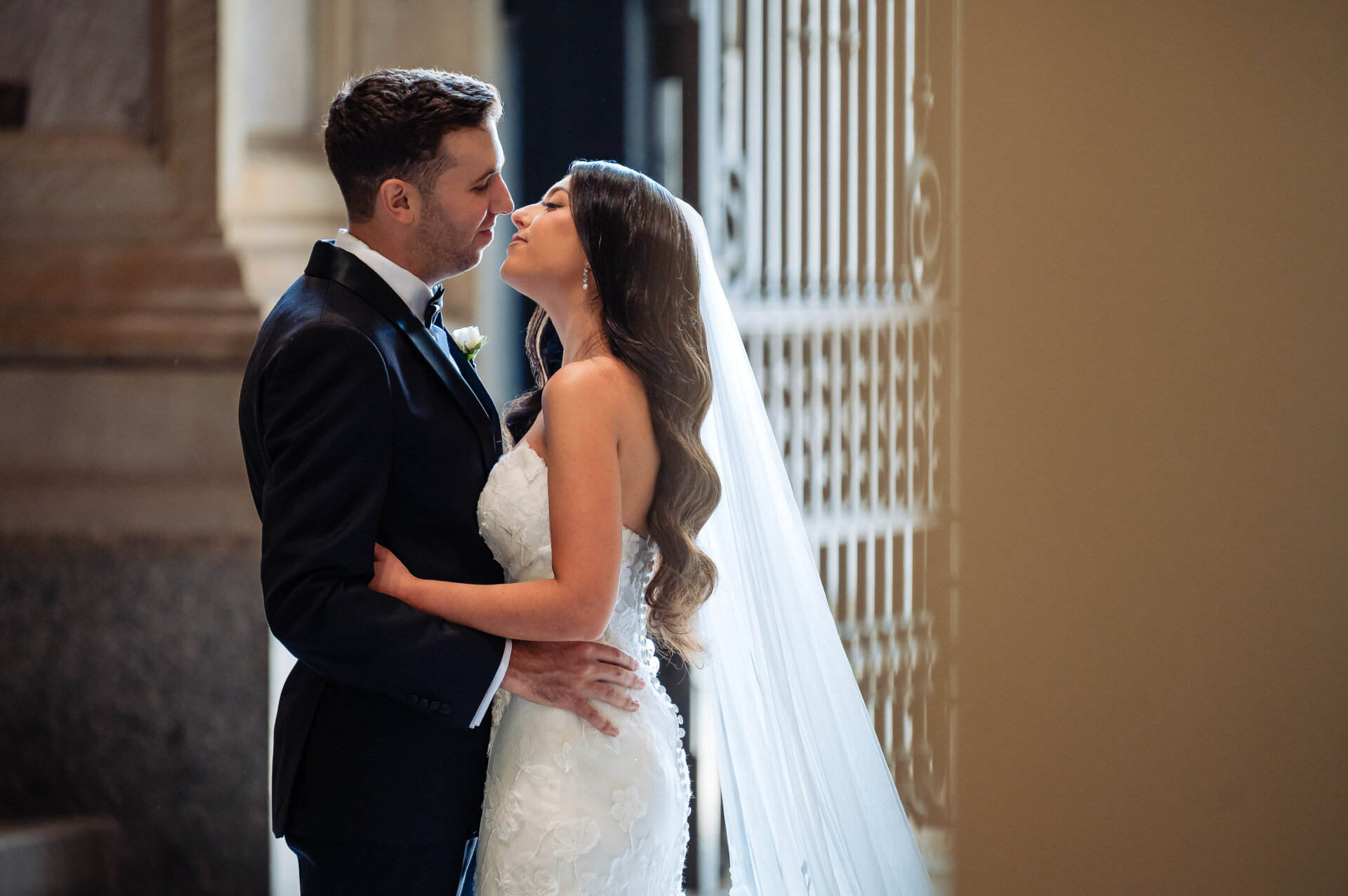 Close-up profile of a bride and groom embracing near a sunlit window, with the bride's long veil catching the light.