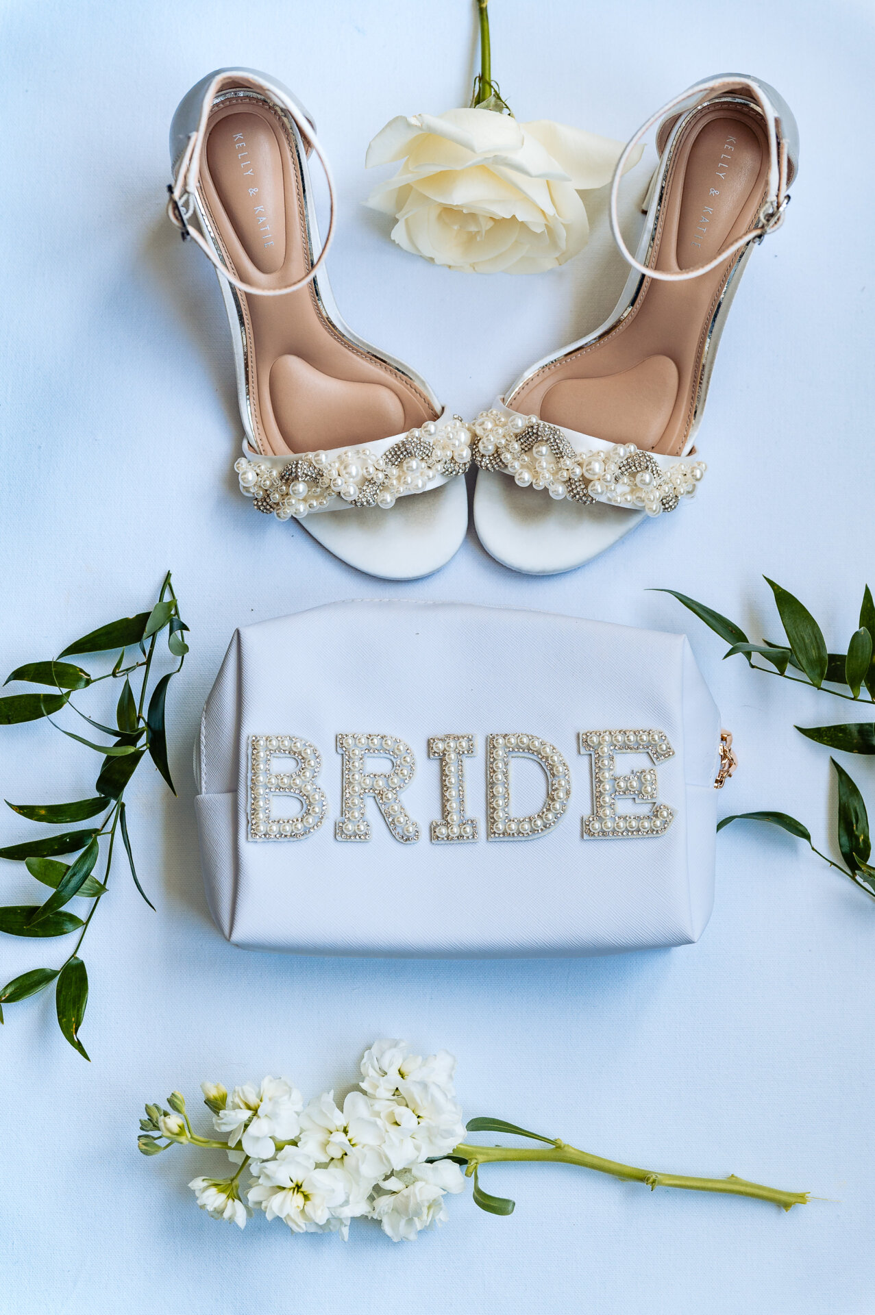 Bridal flatlay with pearl and crystal embellished white wedding heels, white bride clutch with jeweled letters, cream rose, and white flowers on marble background