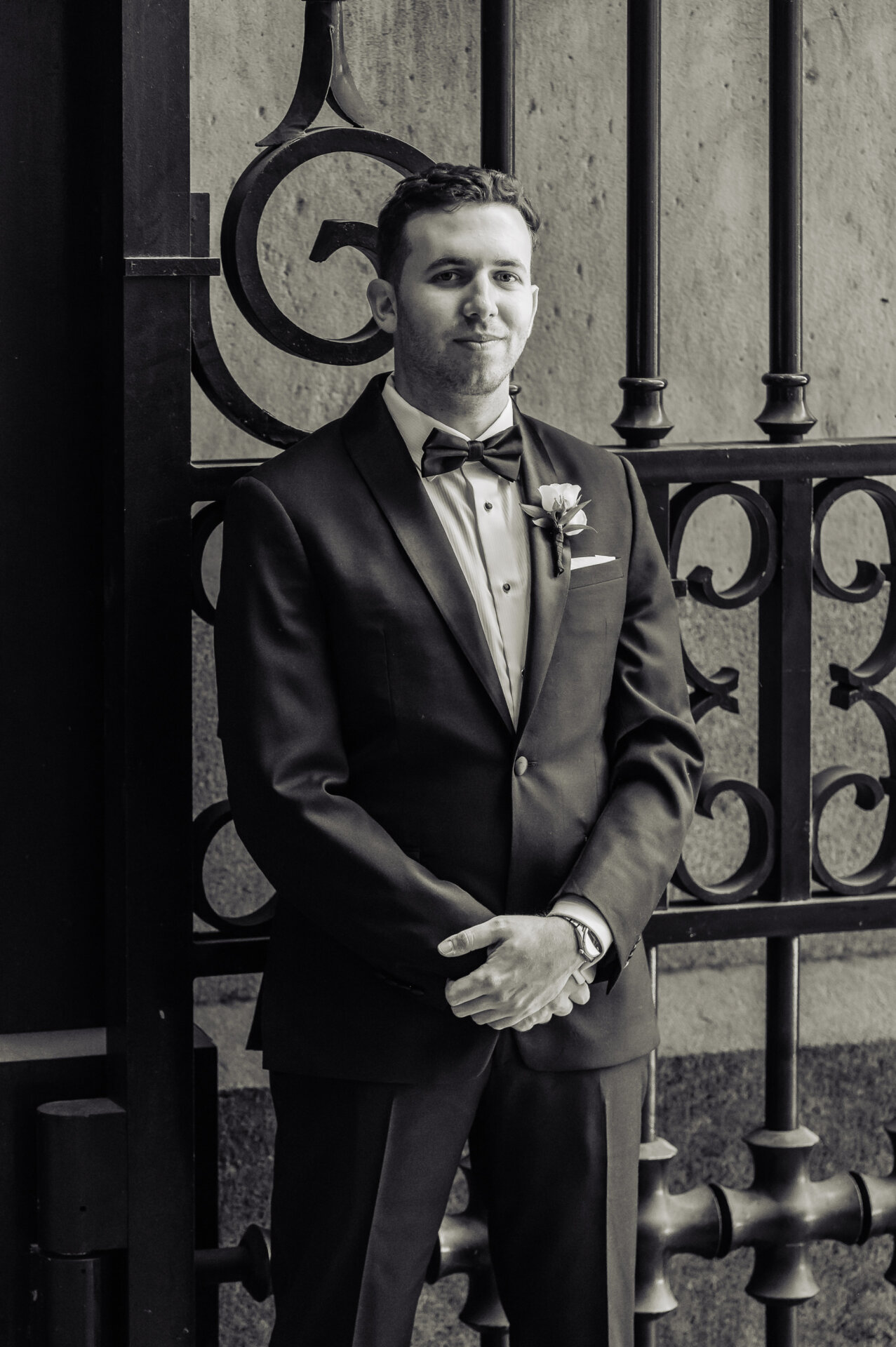 Black and white portrait of groom in tuxedo with bow tie and boutonniere leaning against ornate iron gate
