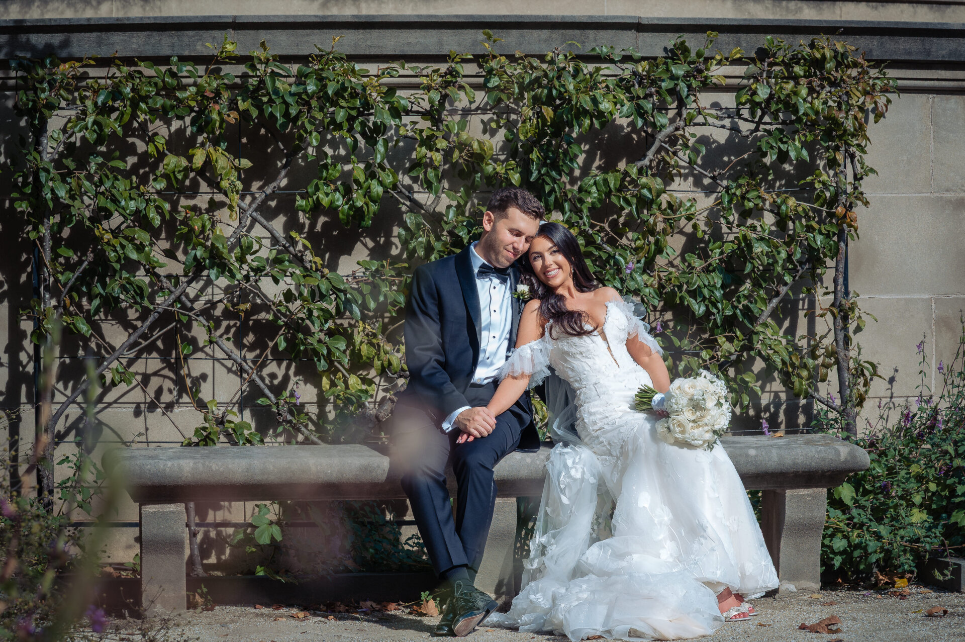 Newlyweds sitting on stone bench against espaliered vine wall, bride holding white bouquet in lace gown with veil