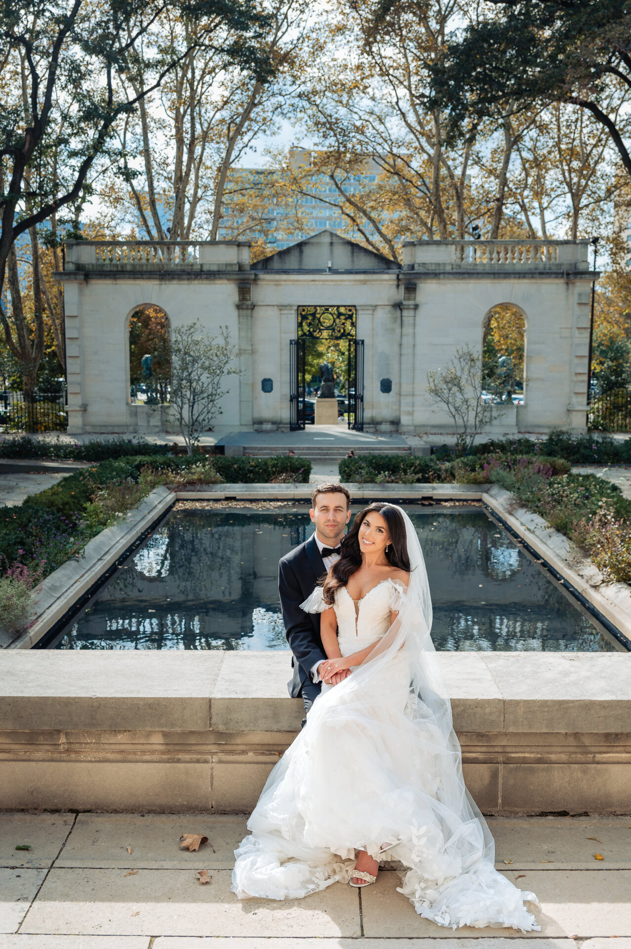Newlyweds sitting on edge of reflecting pool with neoclassical pavilion and autumn trees in background