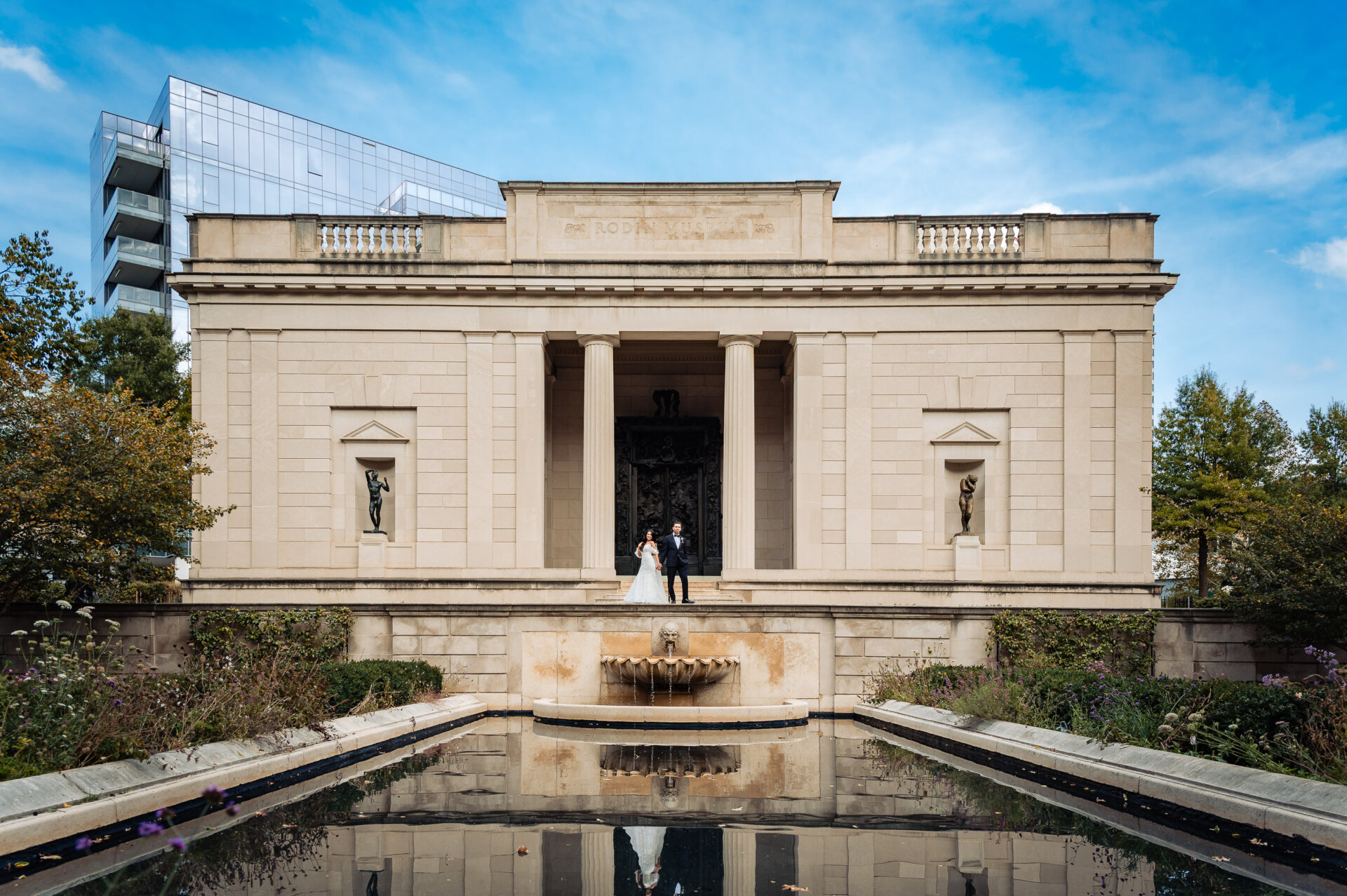 Bride and groom standing on terrace of neoclassical pavilion with reflecting pool and fountain in foreground under blue sky