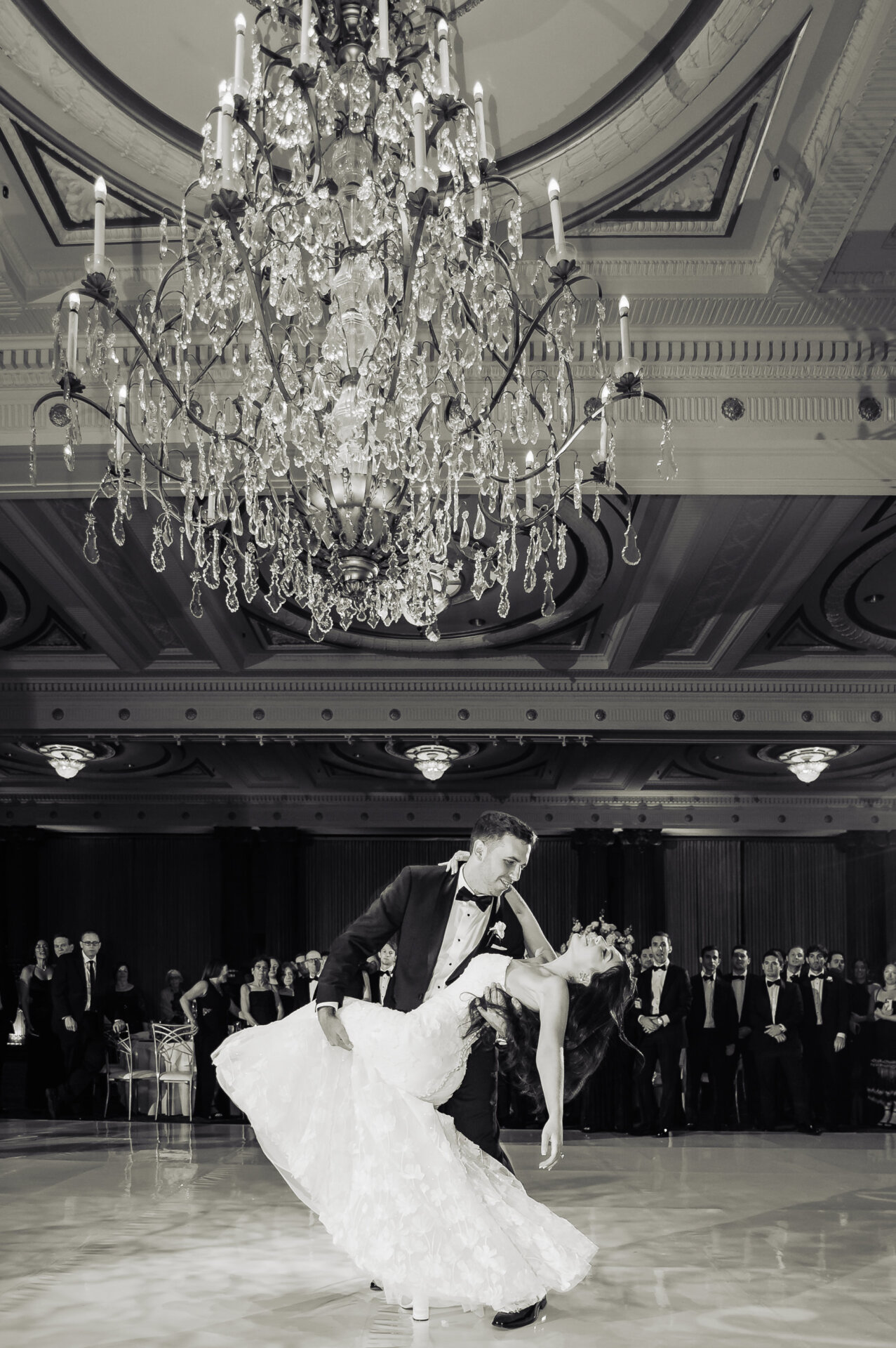 monochromatic shot of the newlywed couple sharing a passionate kiss on a grand ballroom dance floor. The groom in a black tuxedo dips his bride in a white wedding gown with a flowing train and long veil.