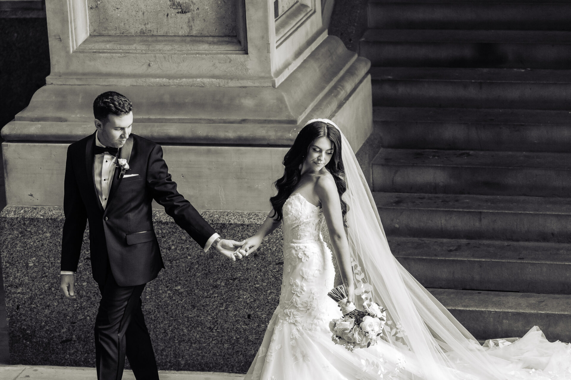 Black and white photo of a bride in a floral lace gown and a groom in a tuxedo walking together and holding hands.