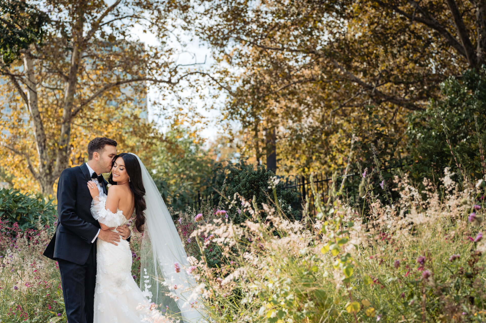Romantic portrait of a groom kissing the bride's forehead in a lush garden filled with wildflowers and autumn trees.