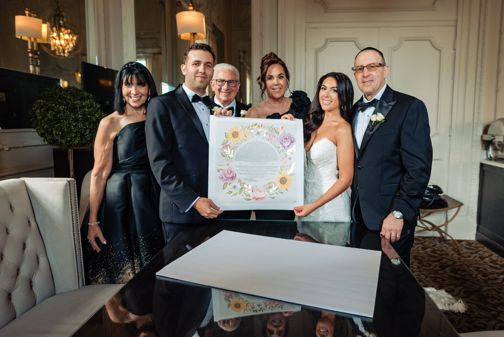 Close-up of a bride and groom looking down at their signed wedding document on a glass table with family standing behind them.