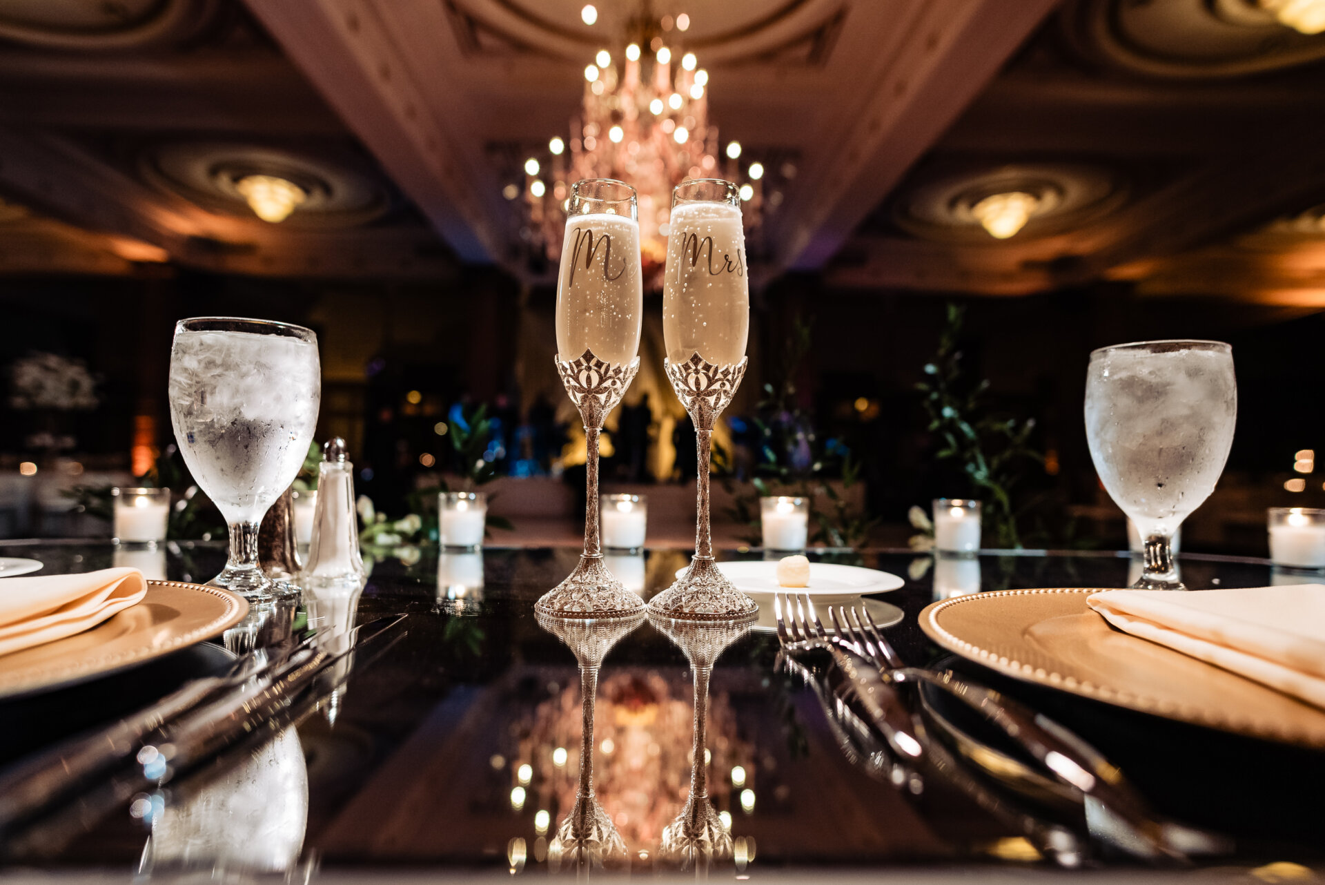 Close-up of engraved Mr & Mrs champagne flutes on a mirrored table with candlelit reception background