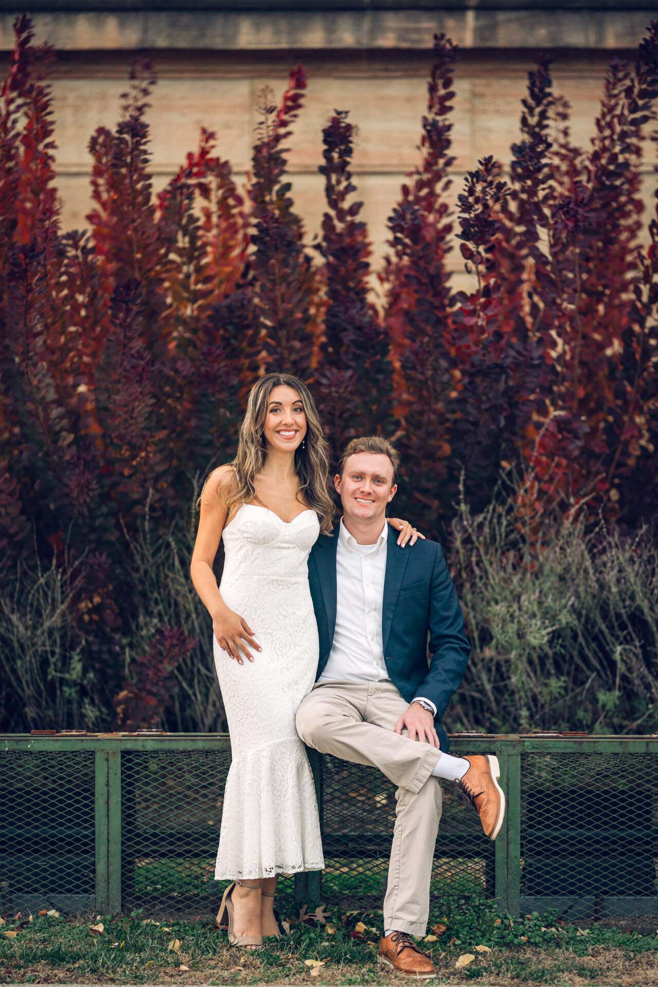 Engaged couple posing together on a bench with fall colors behind them.