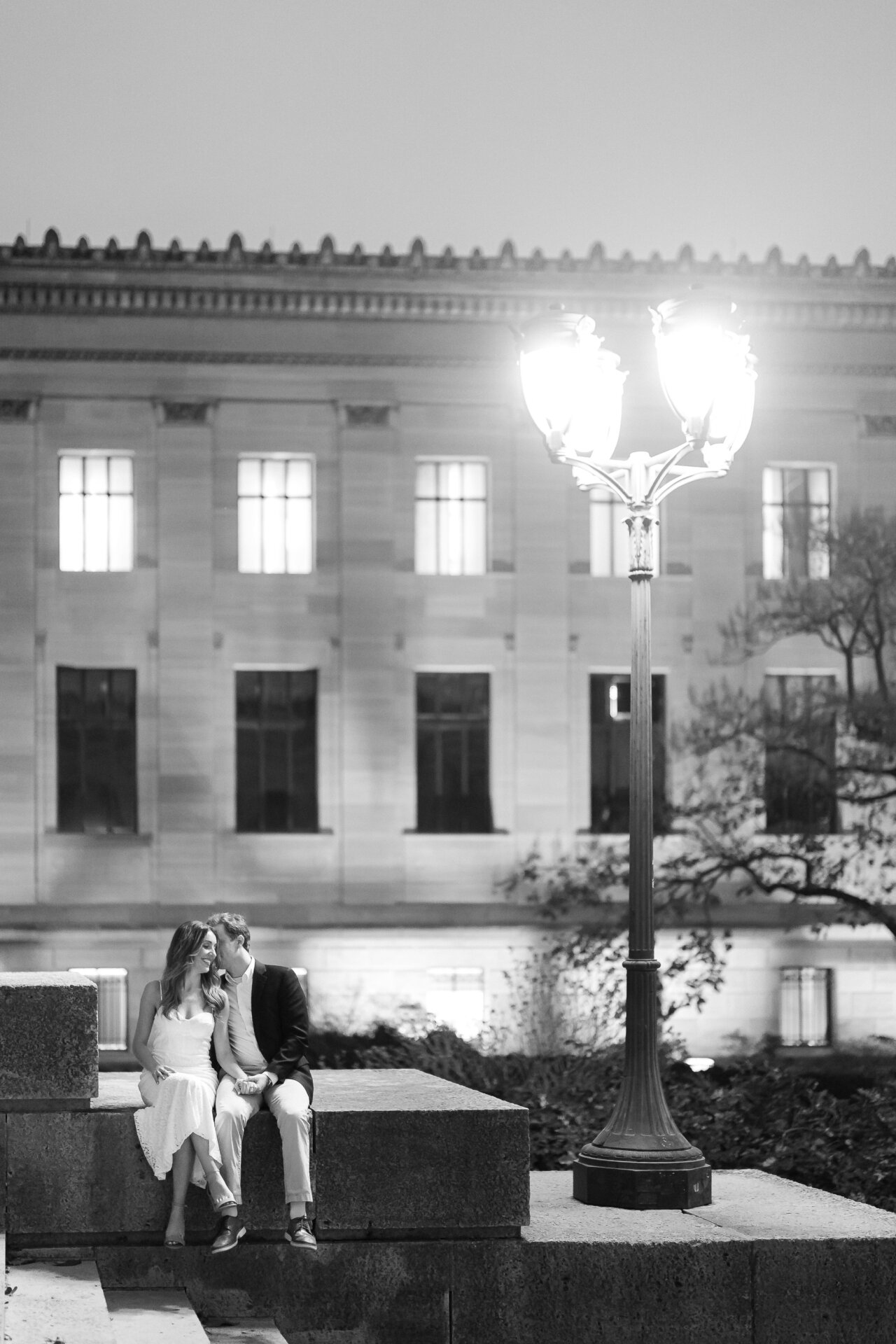 Black-and-white engagement photo of a couple sitting beneath a glowing streetlamp.