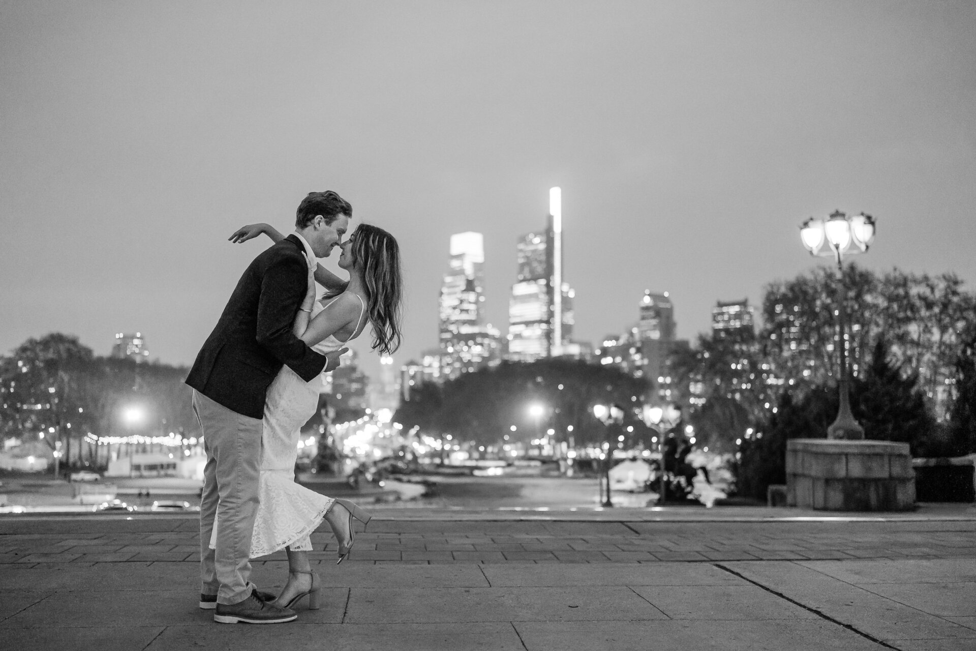 Black-and-white nighttime engagement kiss with the Philadelphia skyline in the background.