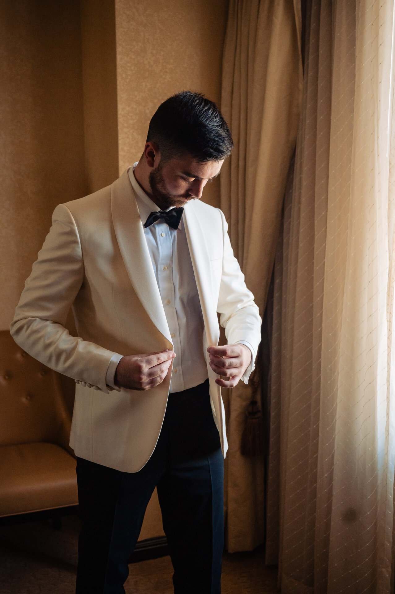 Groom buttoning white dinner jacket with black bow tie looking down in hotel room with natural window light