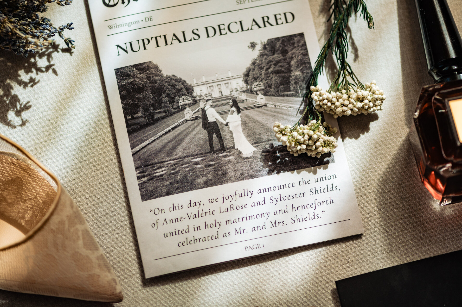 Flat lay of a wedding newspaper announcement, white florals, and perfume arranged on linen fabric.