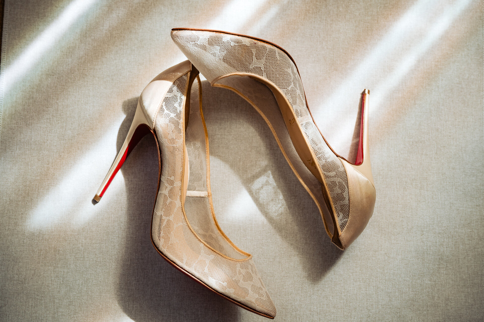 Close-up of ivory lace bridal heels with red soles placed in soft window light.