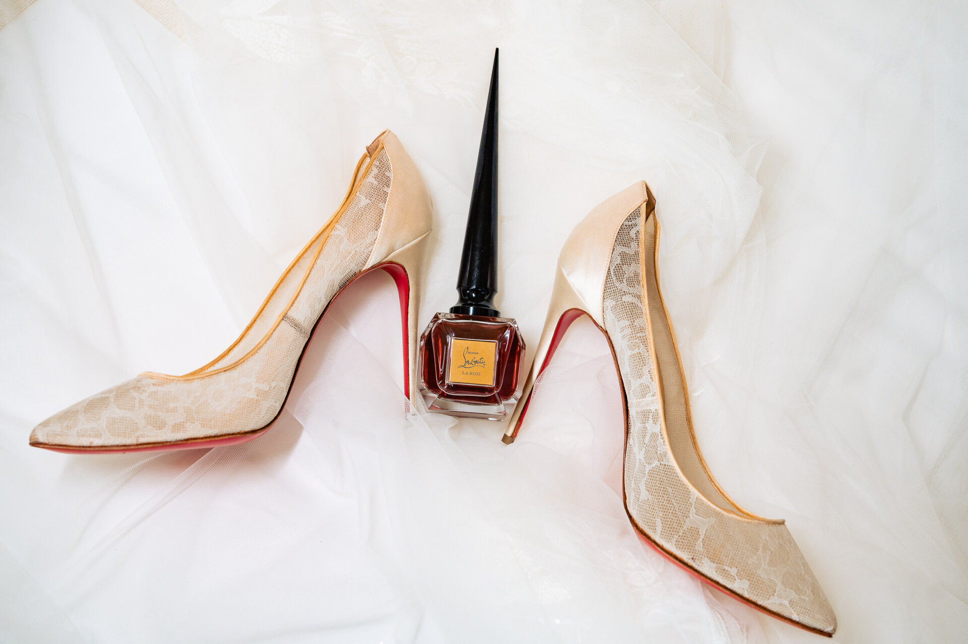 Ivory lace bridal heels with red soles placed beside a bottle of perfume on white fabric.