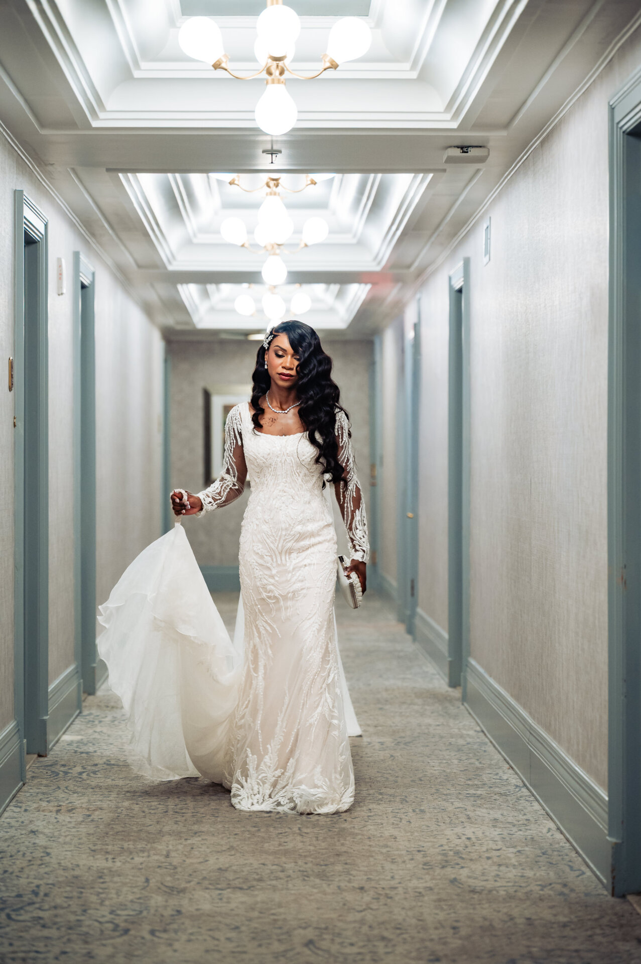 Bride in white lace mermaid gown with long veil walking down modern hotel hallway with coffered ceiling