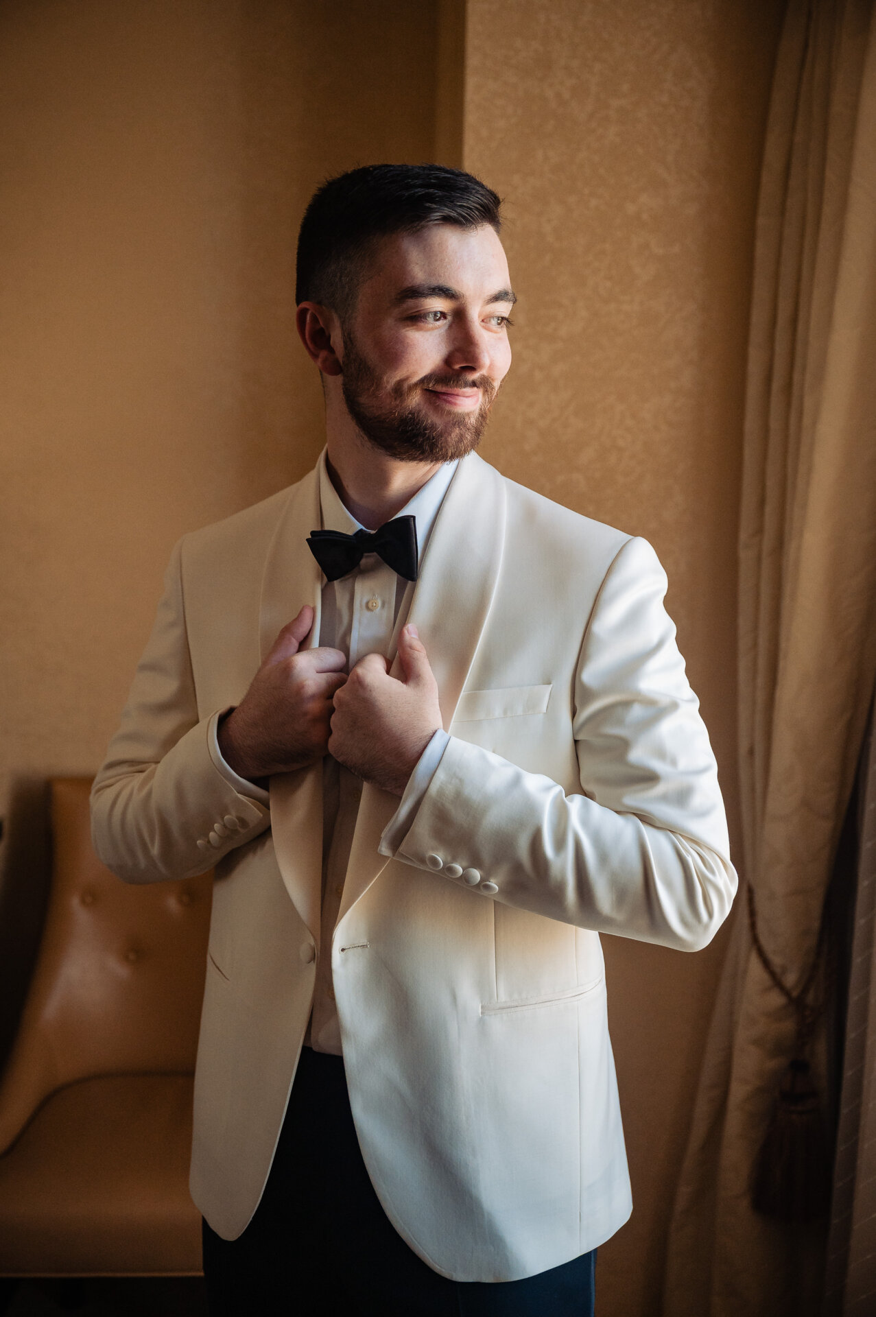 Groom adjusting white dinner jacket with black bow tie in warm-lit hotel room