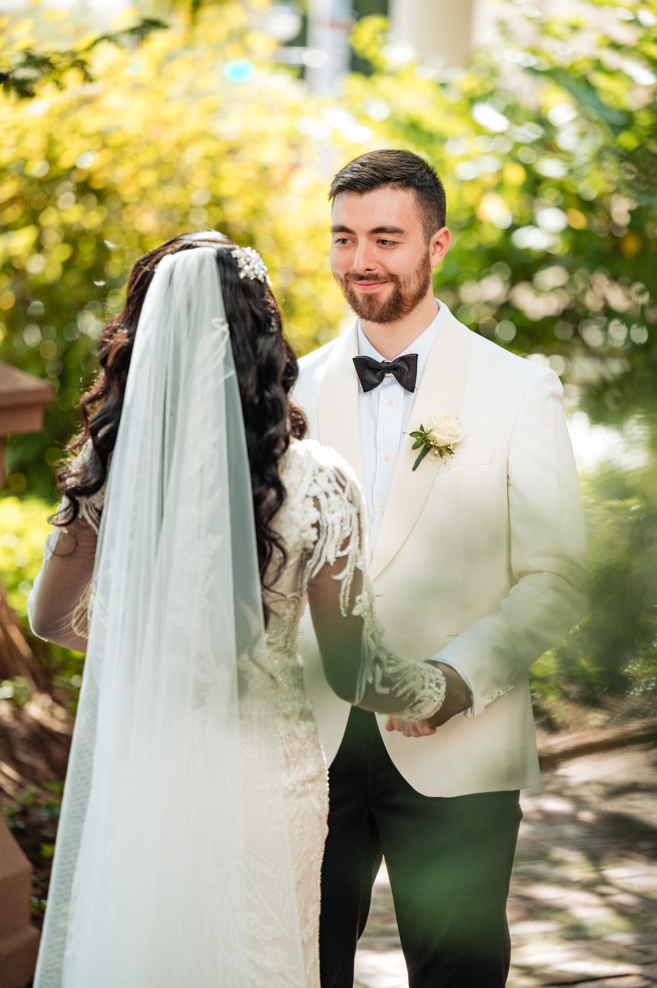 Groom in white dinner jacket smiling at bride during first look in sunlit garden with bokeh foliage
