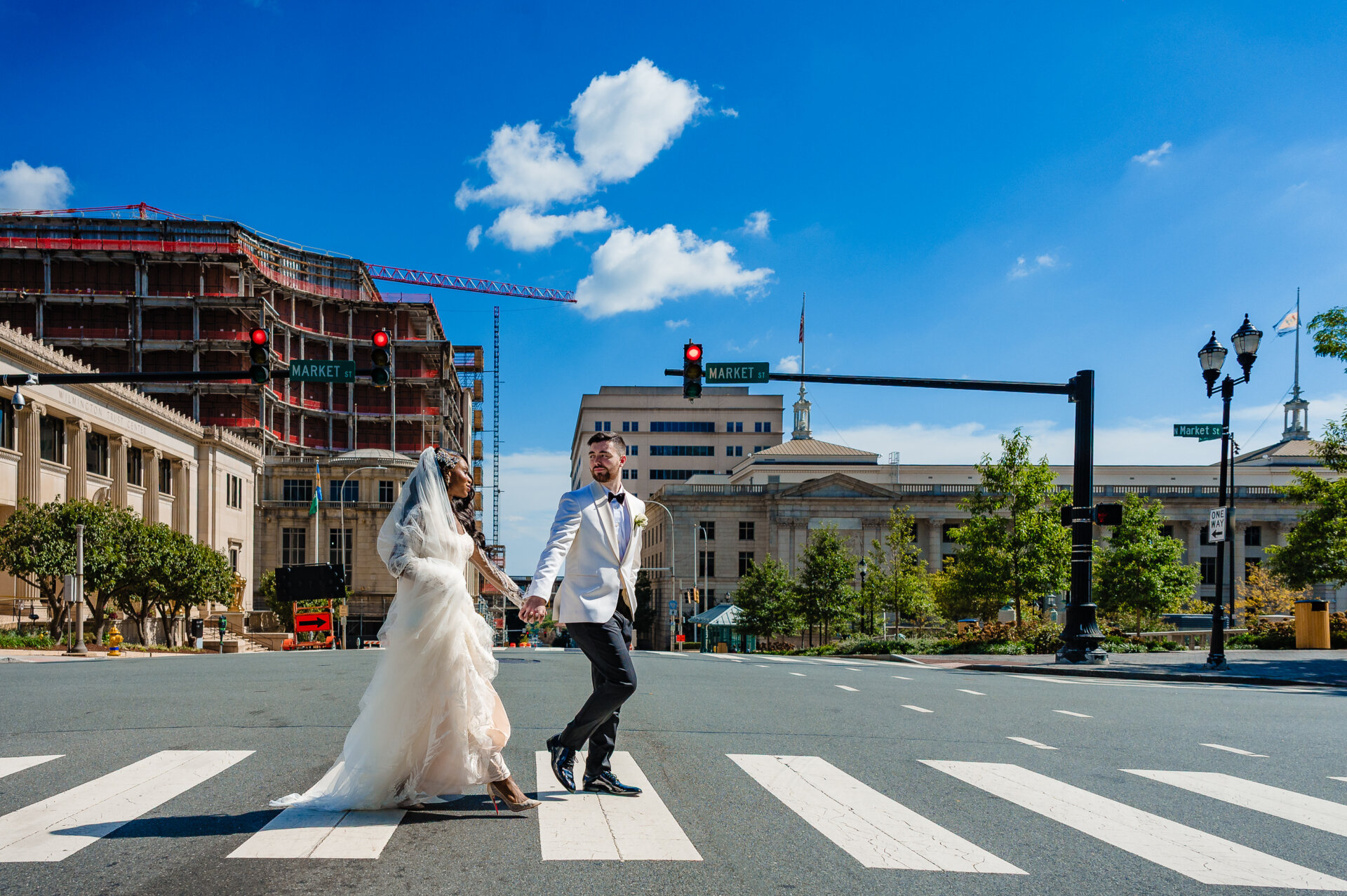 Bride and groom holding hands while crossing urban crosswalk with city buildings and blue sky in background