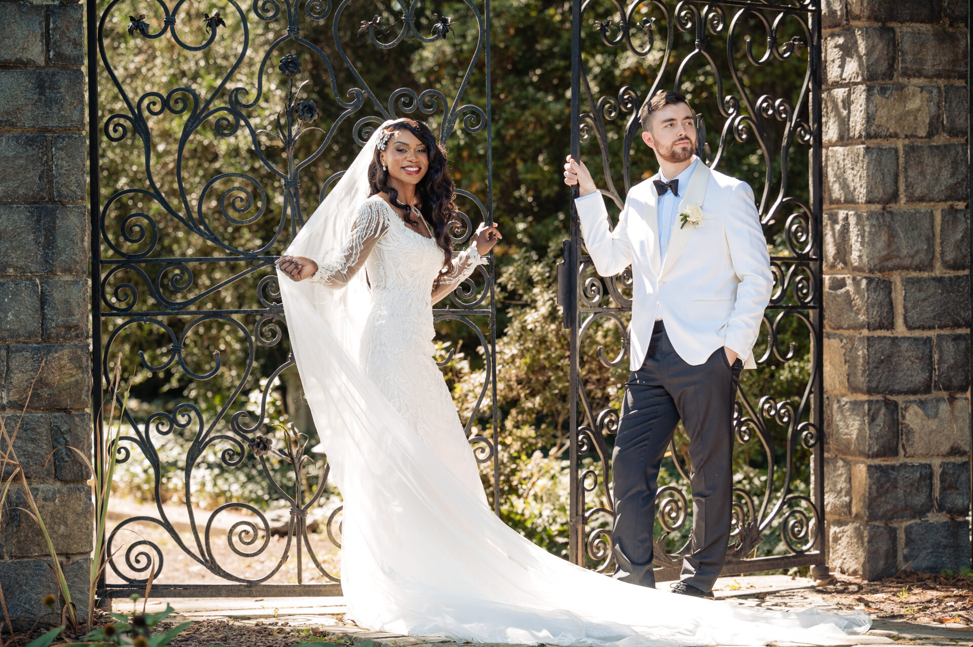 Bride and groom walking through ornate iron gate with ivy-covered stone pillars in bright sunlight