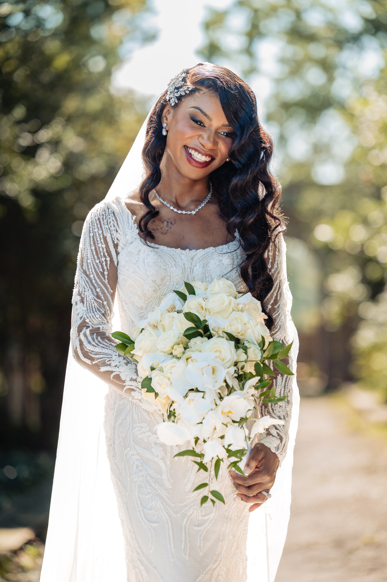 Bride smiling and holding white and green cascading bouquet in long-sleeve lace gown with natural bokeh background