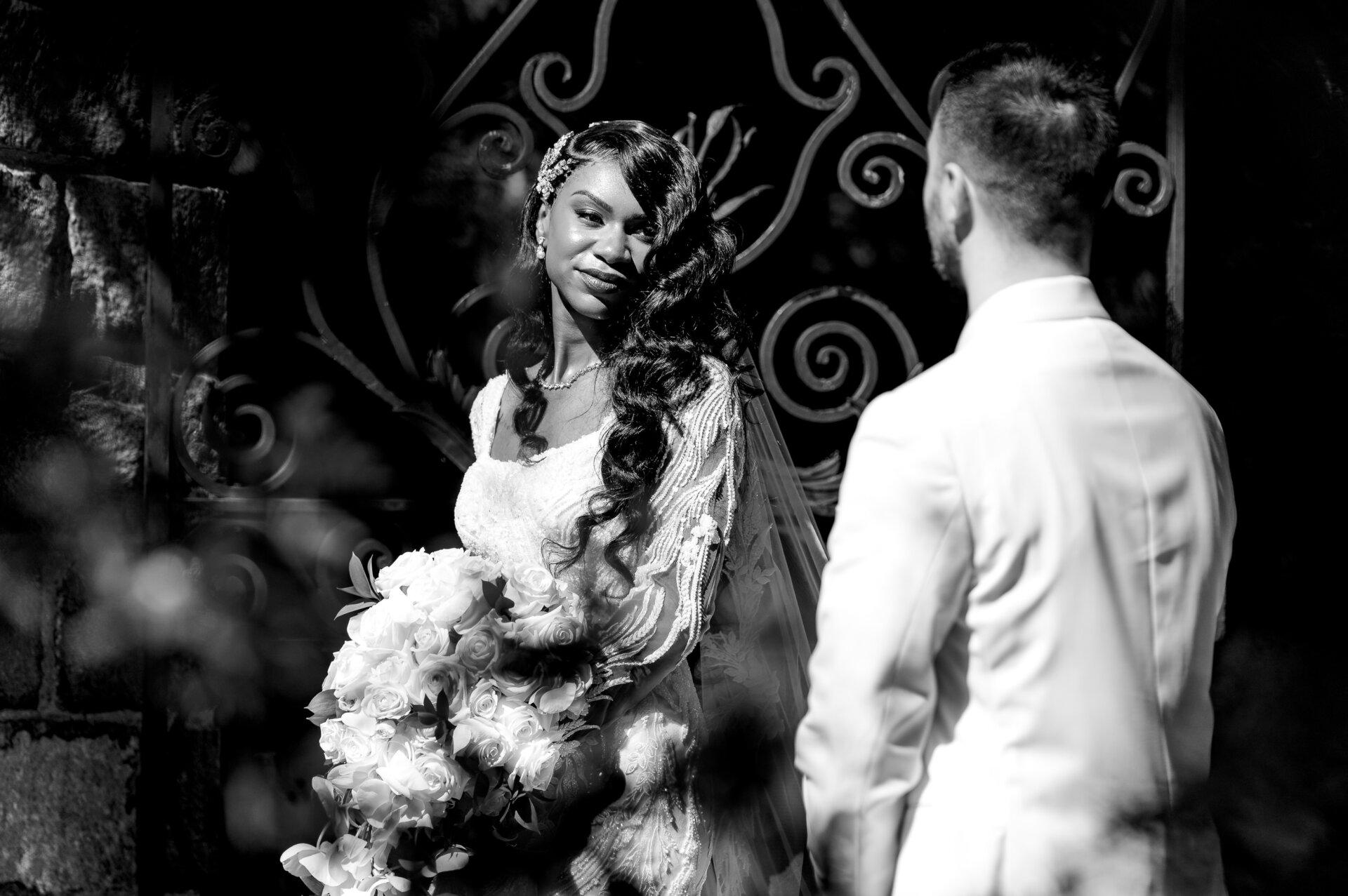 Black and white photo of bride and groom facing each other with white bouquet against decorative iron scrollwork