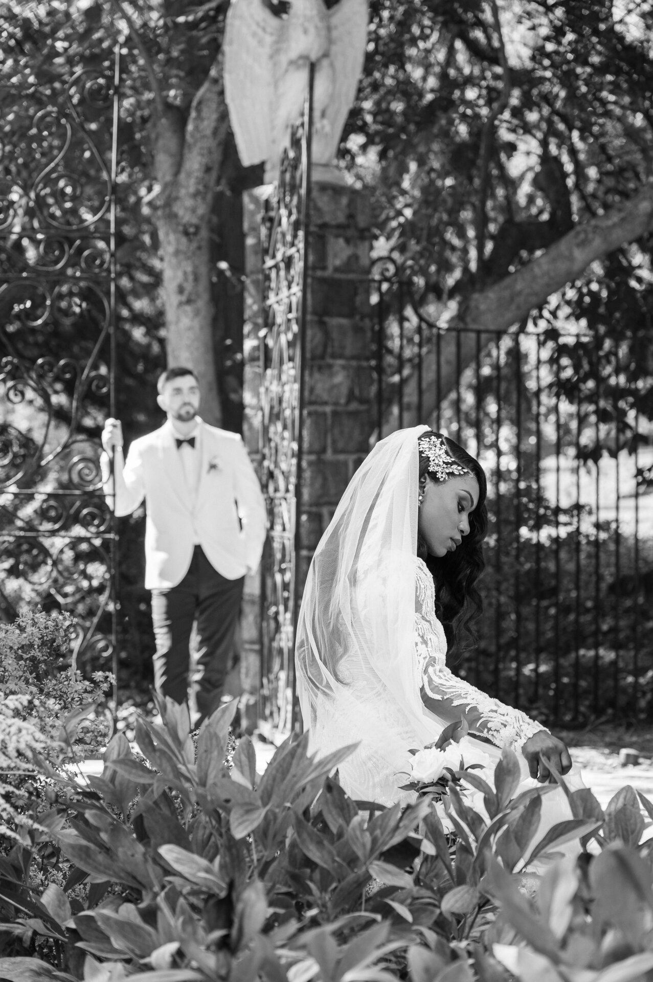Black and white photo of a bride adjusting her veil in a garden while the groom approaches from behind.