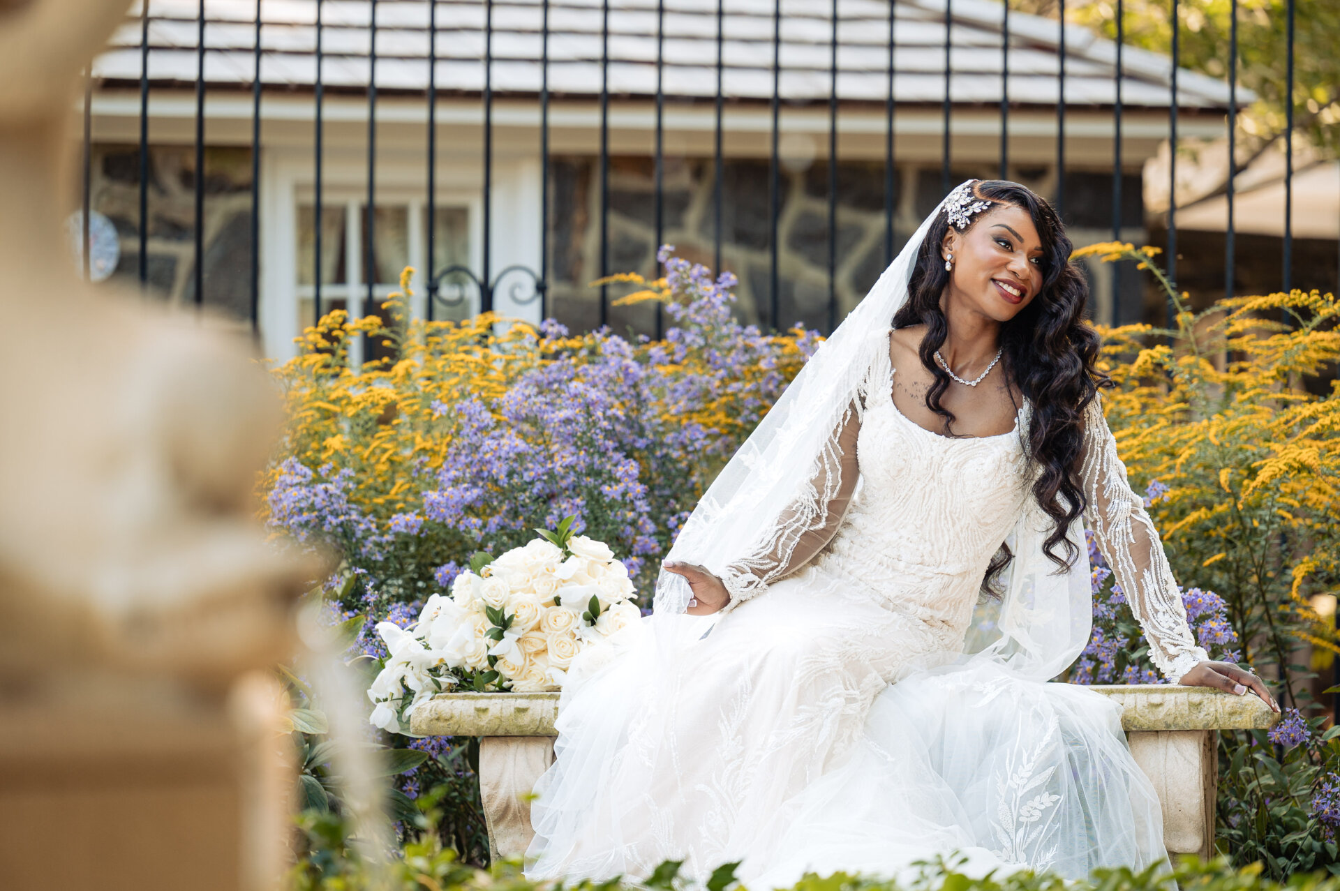 Bride seated on a stone bench in a garden holding her bouquet, surrounded by blooming flowers.