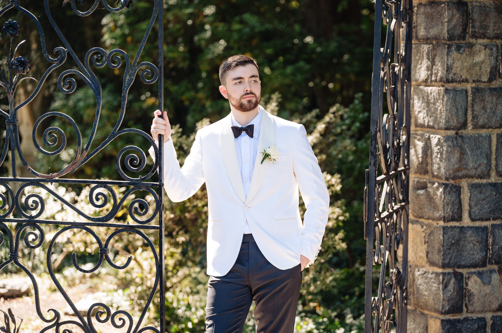 Groom standing alone at iron gates wearing a white dinner jacket and black bow tie.