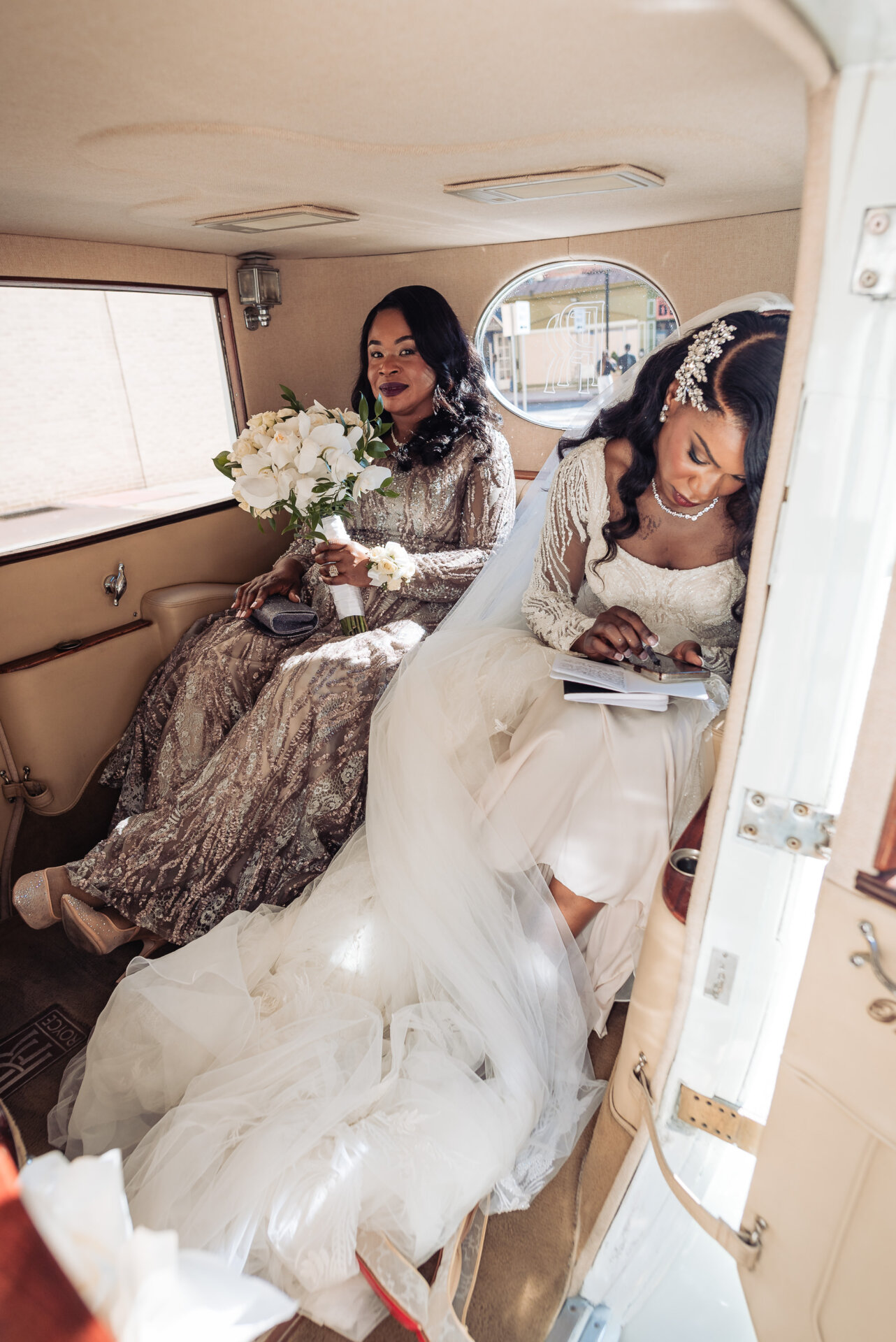 Bride sitting in a vintage car holding her bouquet while a bridesmaid smiles beside her.