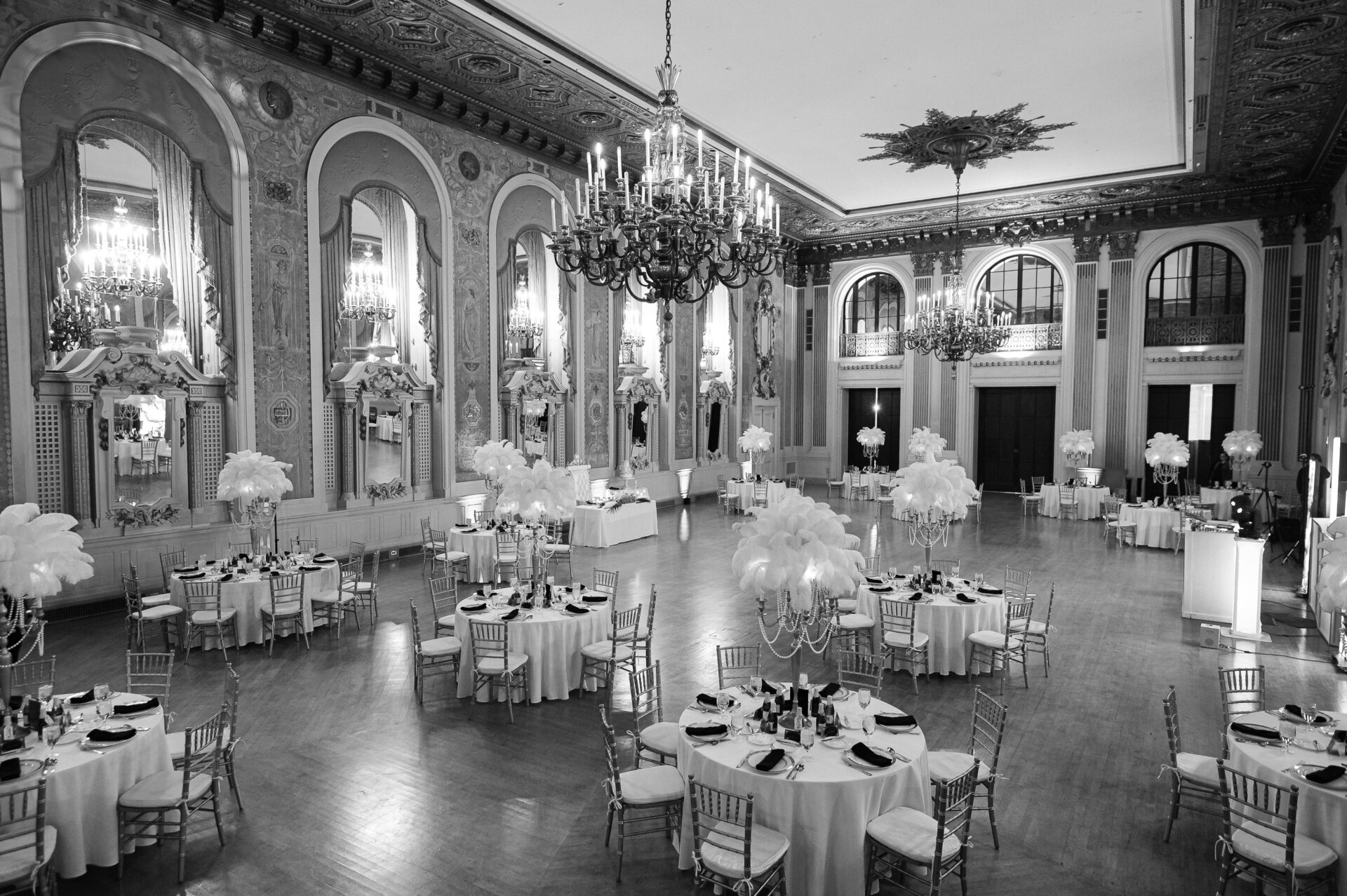 Black and white view of a grand ballroom reception setup with round tables, chandeliers, and feather centrepieces.