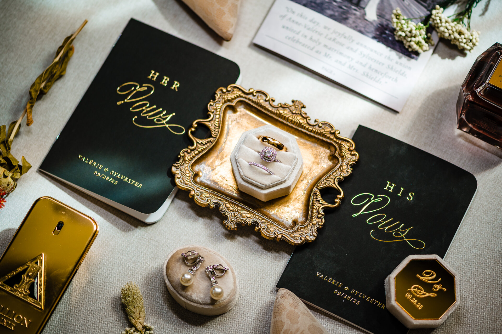 Flat lay of wedding details including vow books, rings in a decorative box, earrings, perfume, and printed stationery.