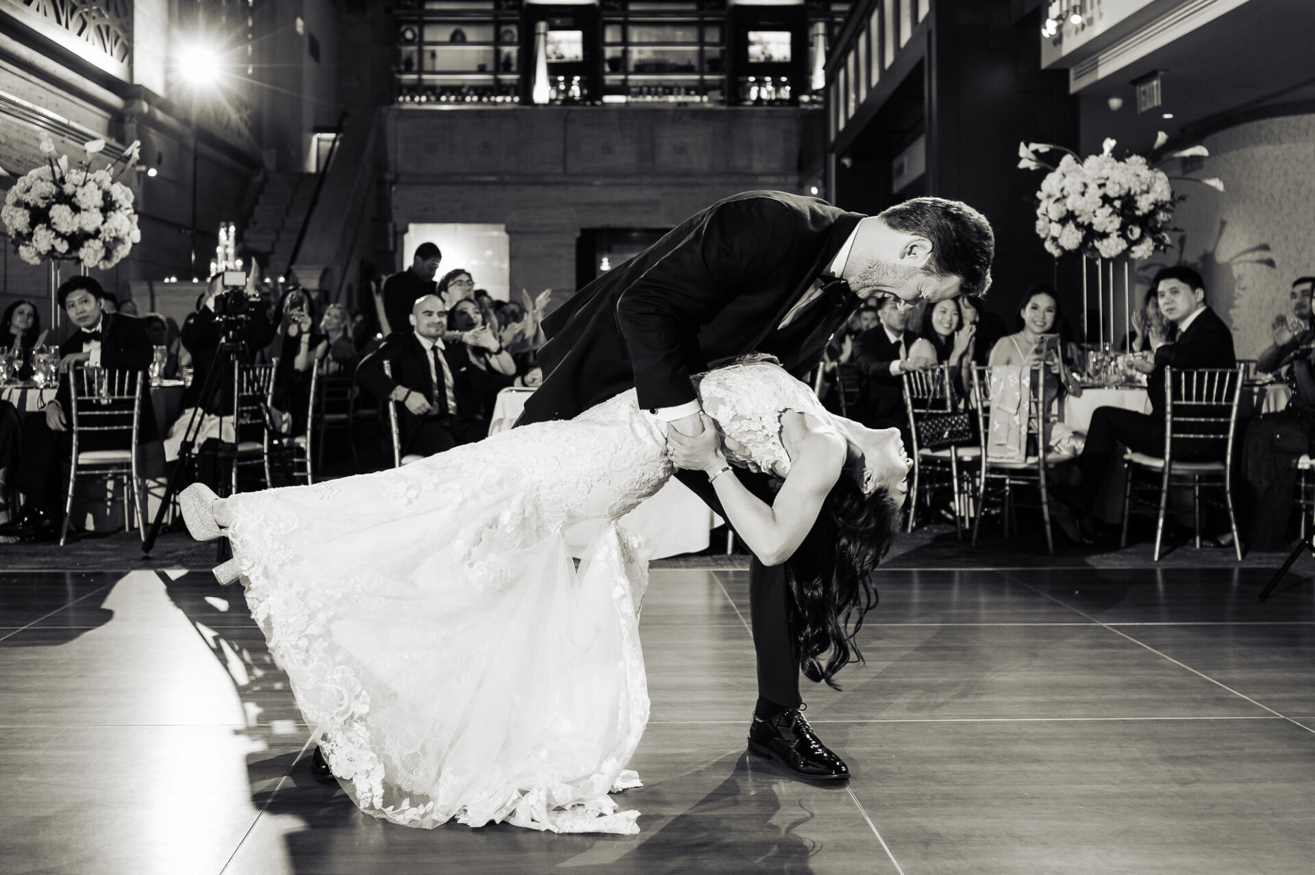 Dramatic black-and-white first dance dip surrounded by cheering wedding guests
