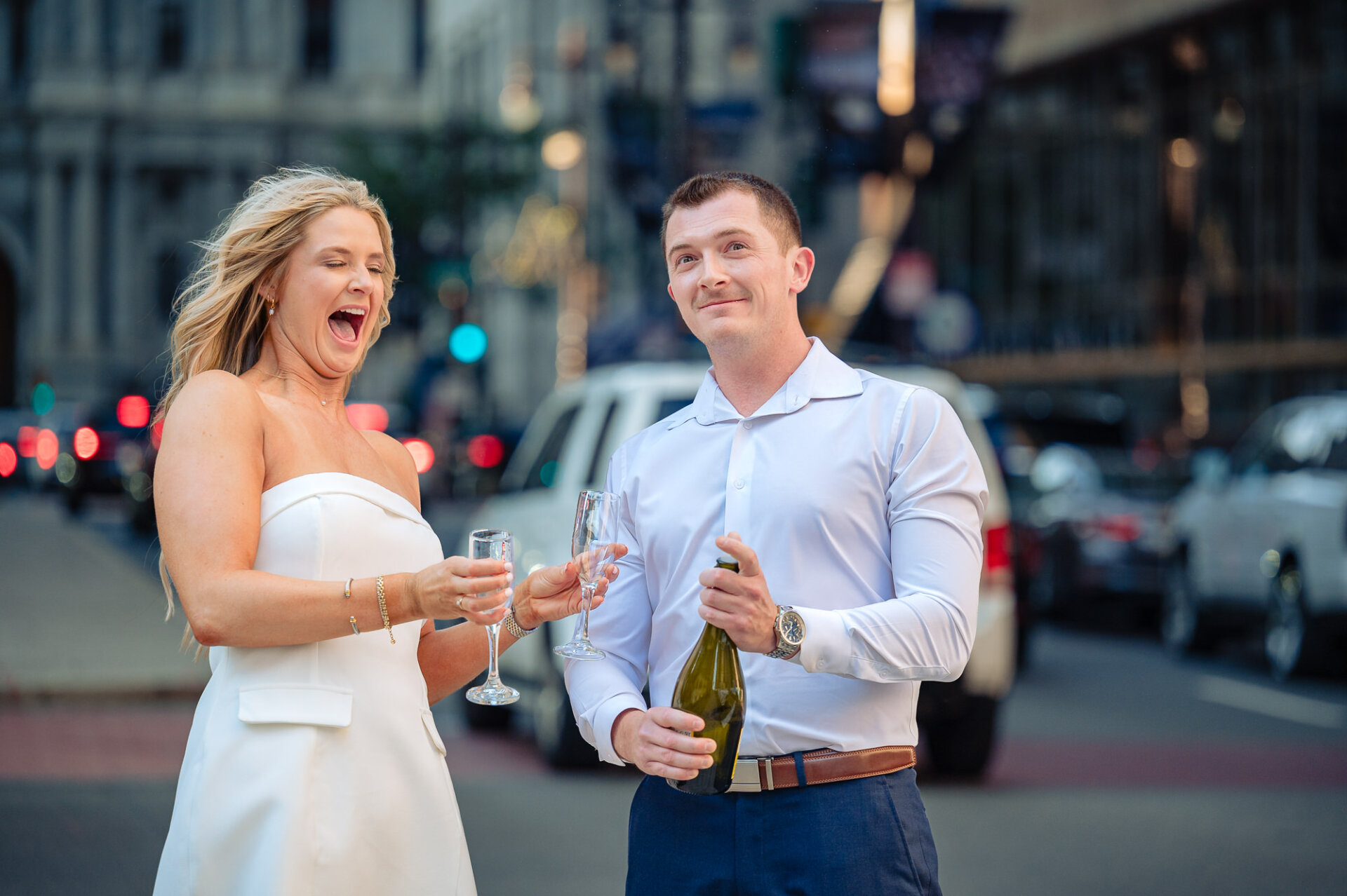 Groom popping champagne with a high foam spray as bride laughs during Center City Philadelphia engagement session