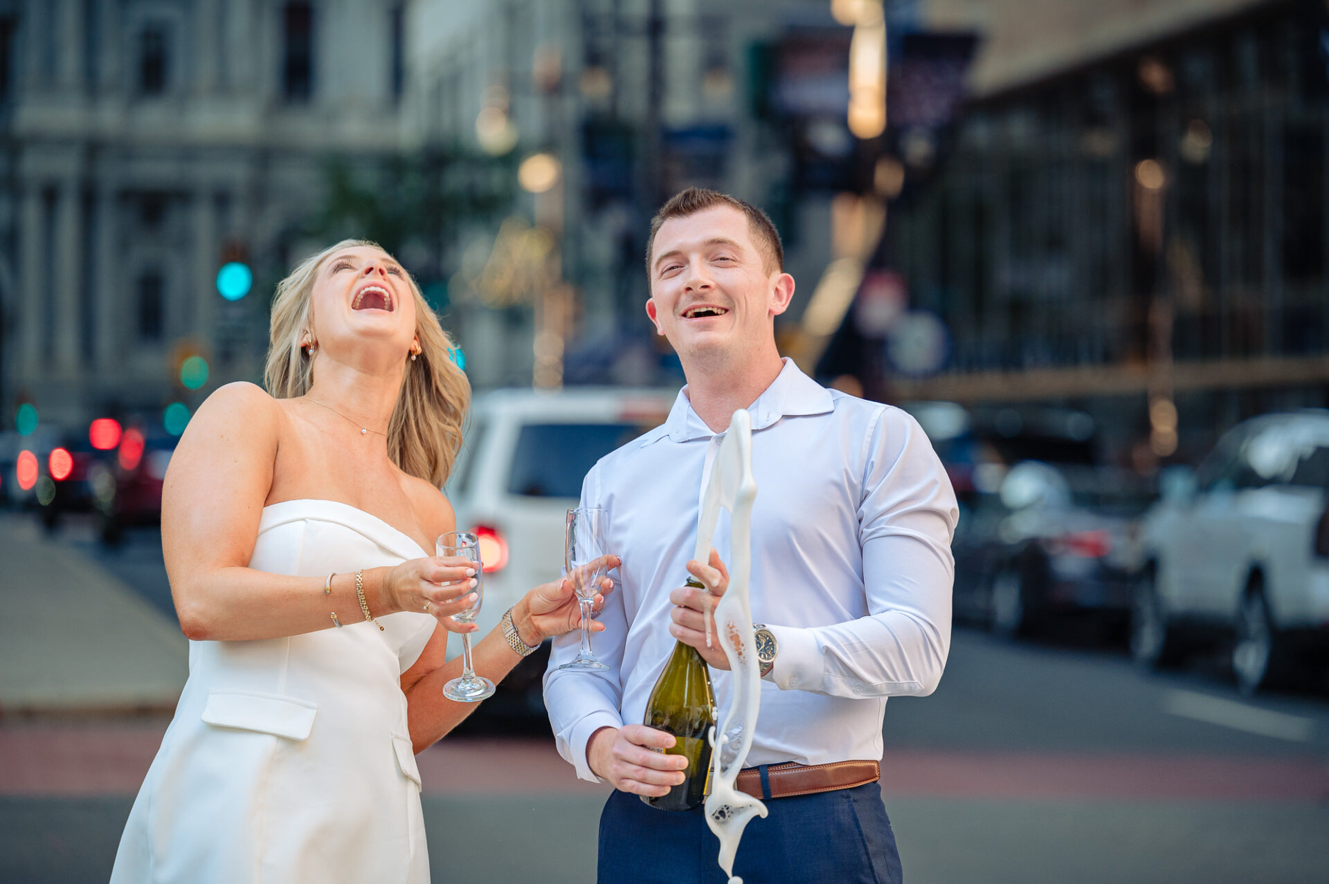 Groom popping champagne with a high foam spray as bride laughs during Center City Philadelphia engagement session
