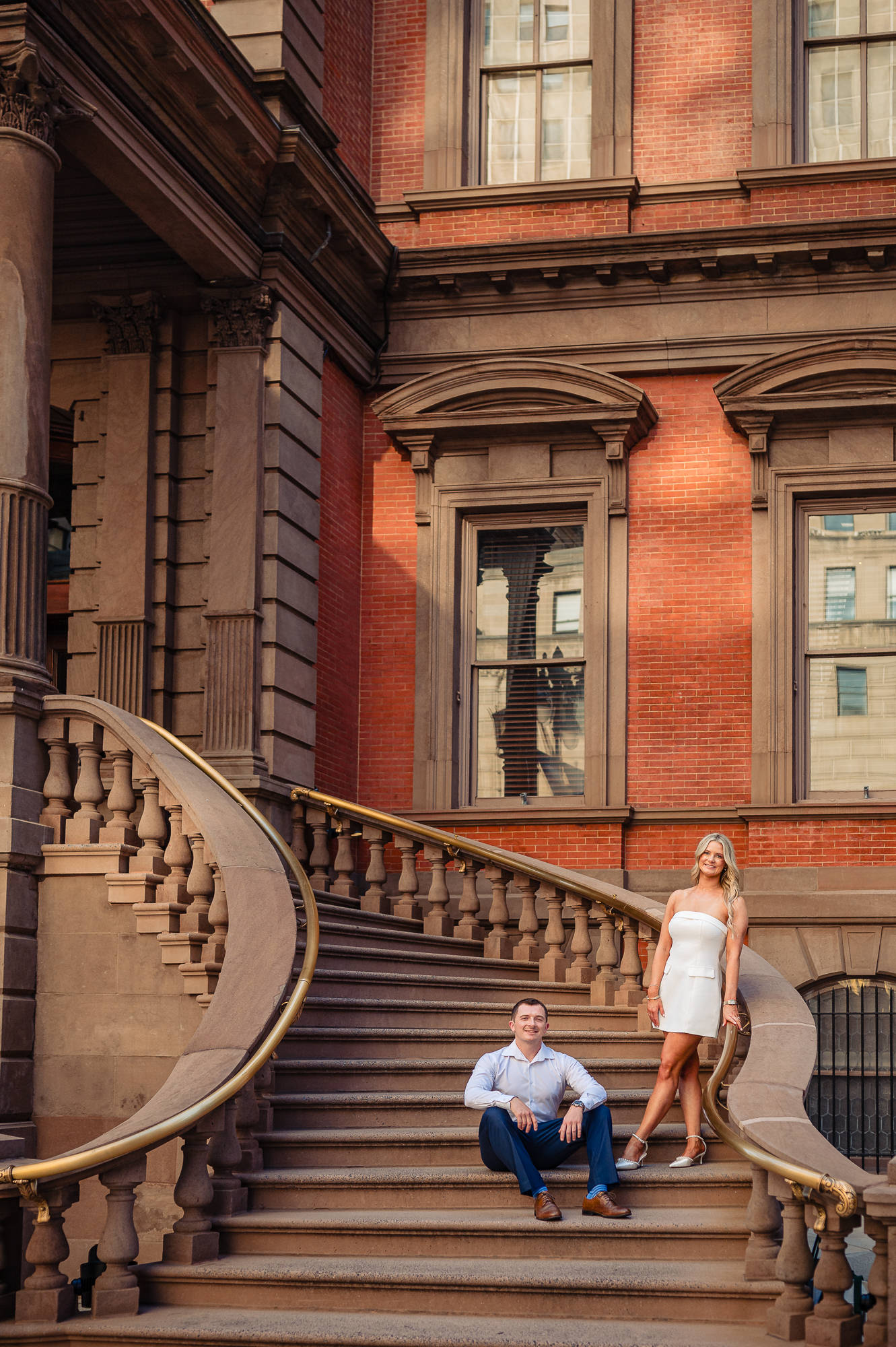 Groom sitting on stone steps while bride stands beside him on the grand staircase of the Union League building in Philadelphia