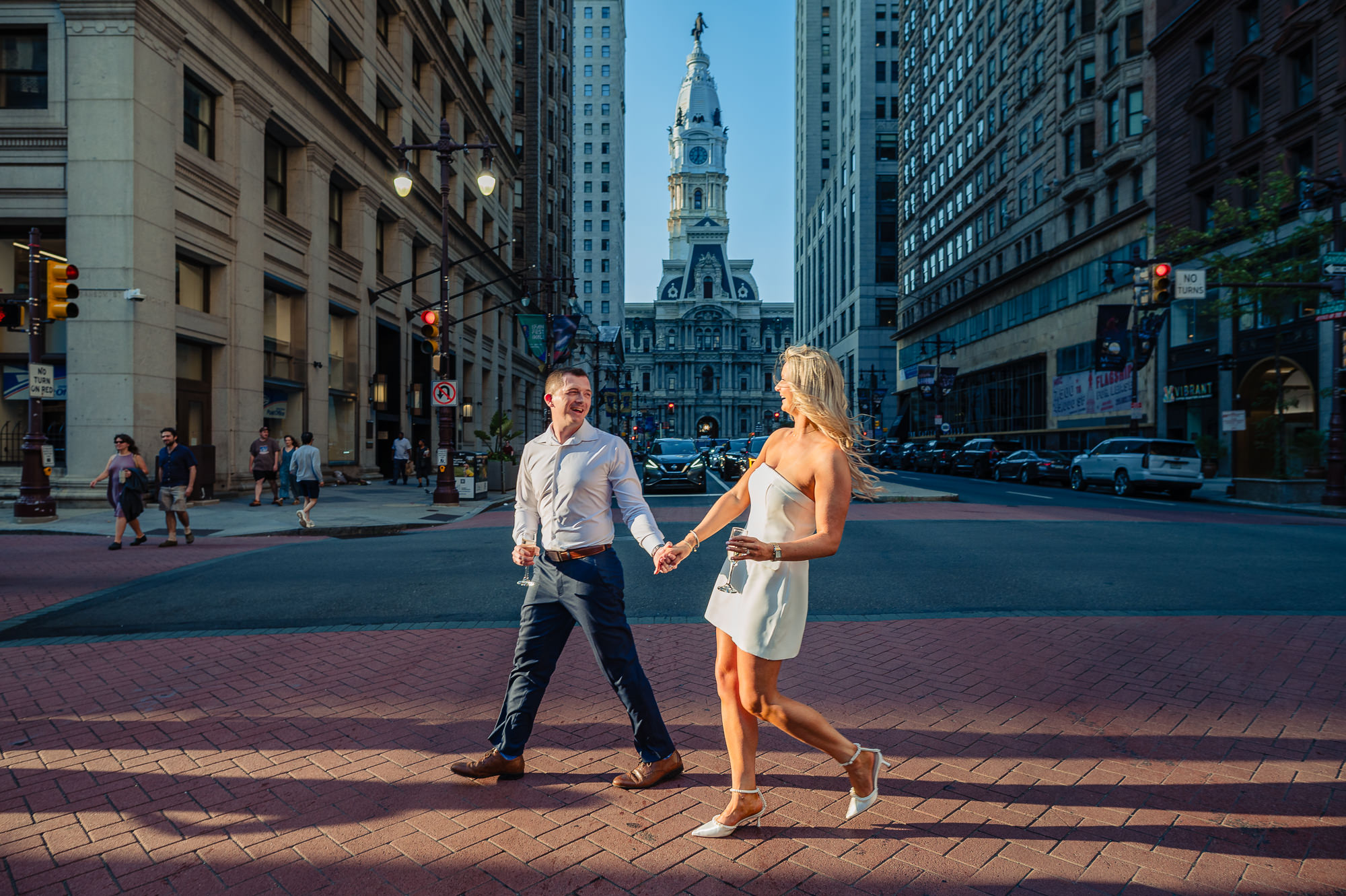Wide shot of engaged couple holding champagne glasses and laughing while walking across Broad Street with City Hall behind them